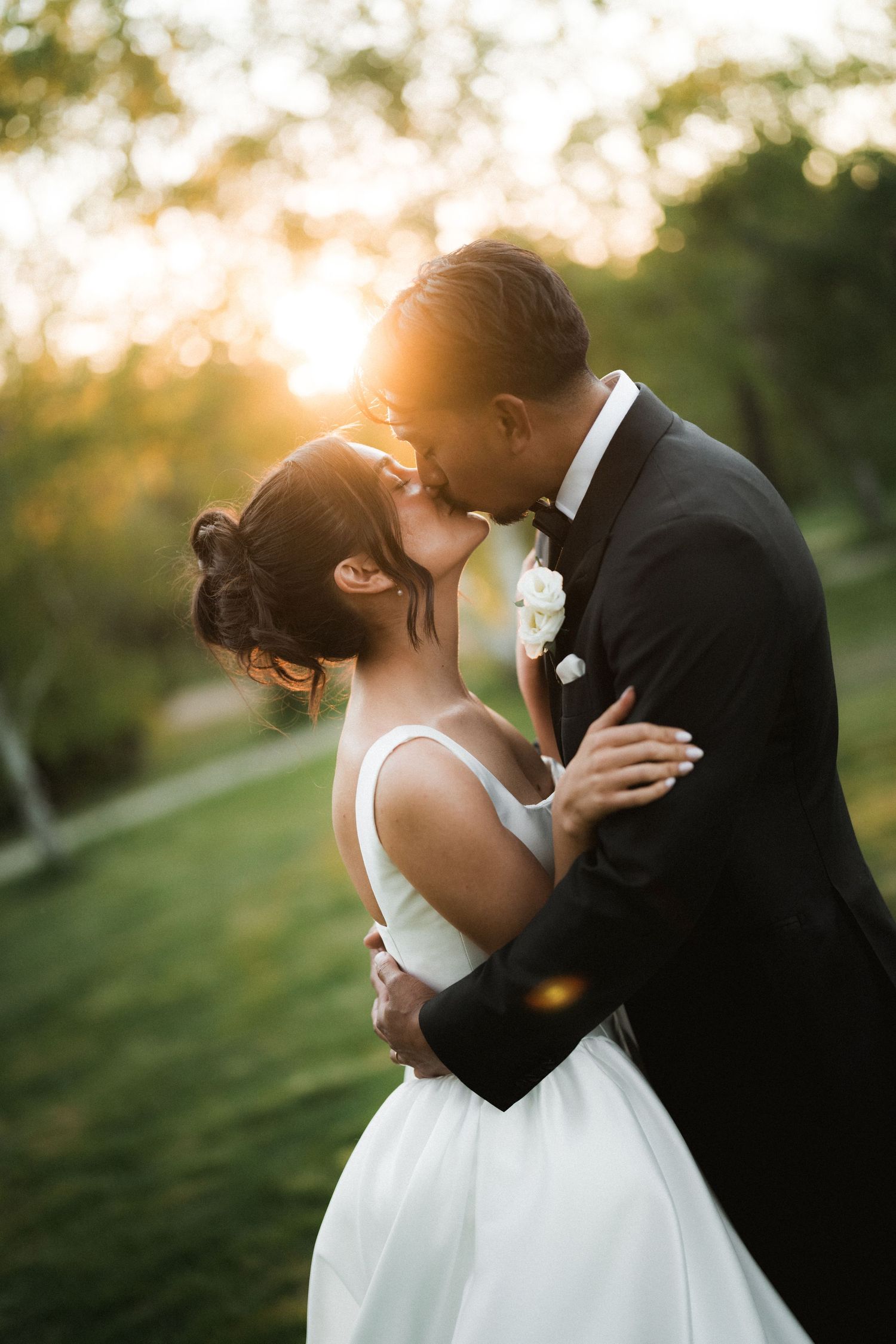 Bride in a white gown and groom in a black tuxedo share a kiss on a grassy lawn at golden hour sunset.