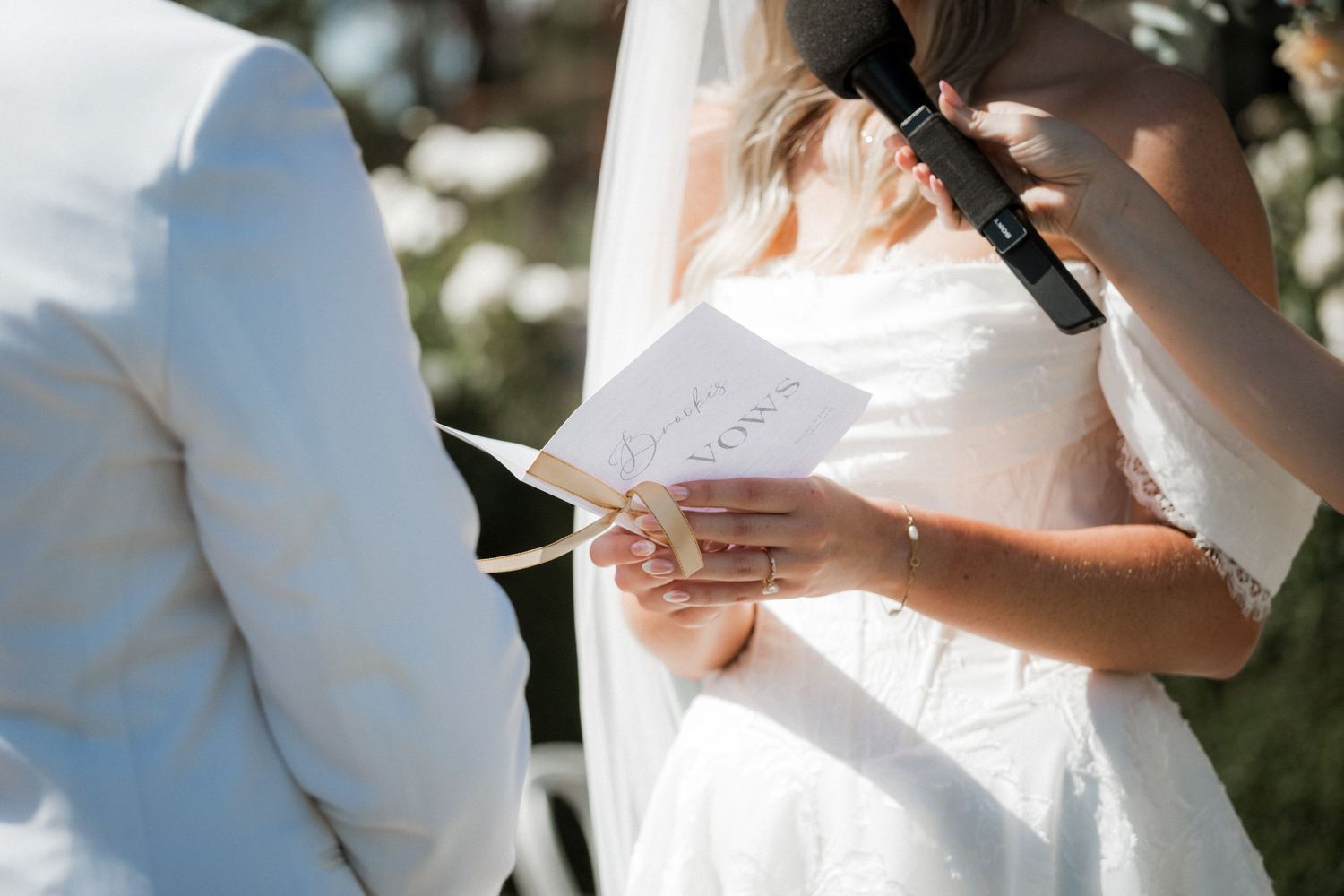Bride reading vows from card at outdoor wedding ceremony holding microphone.