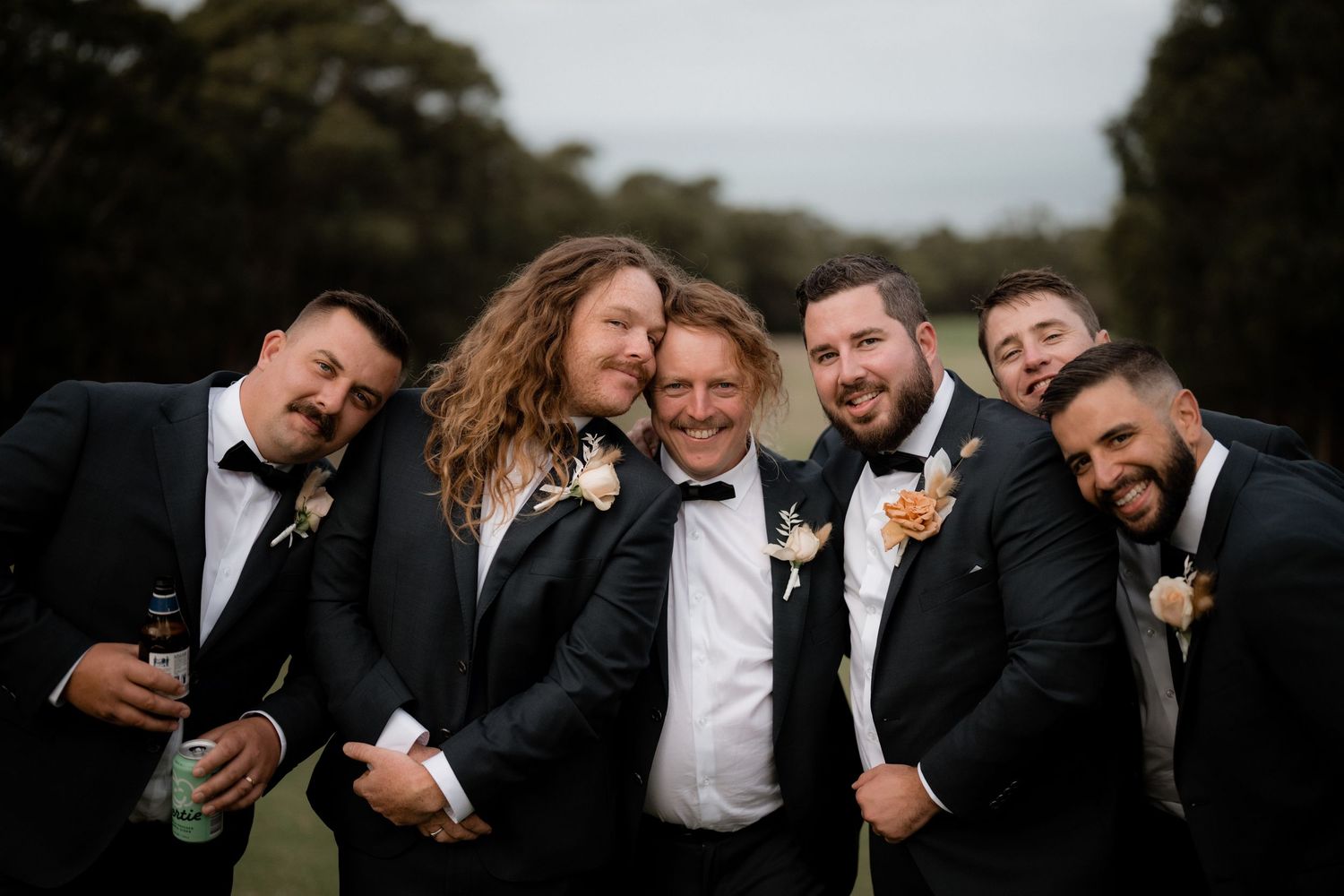 A wedding party in black tuxedos gathers closely for a candid group photo outdoors.