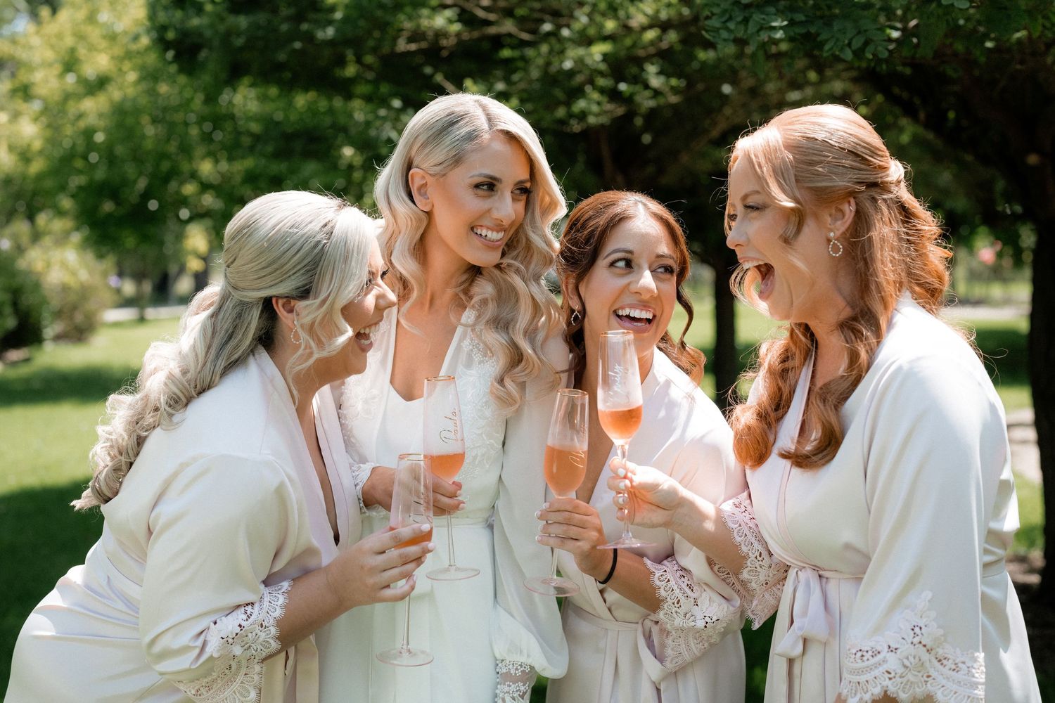 Four women in white robes laugh and celebrate together while holding champagne glasses in a garden setting.