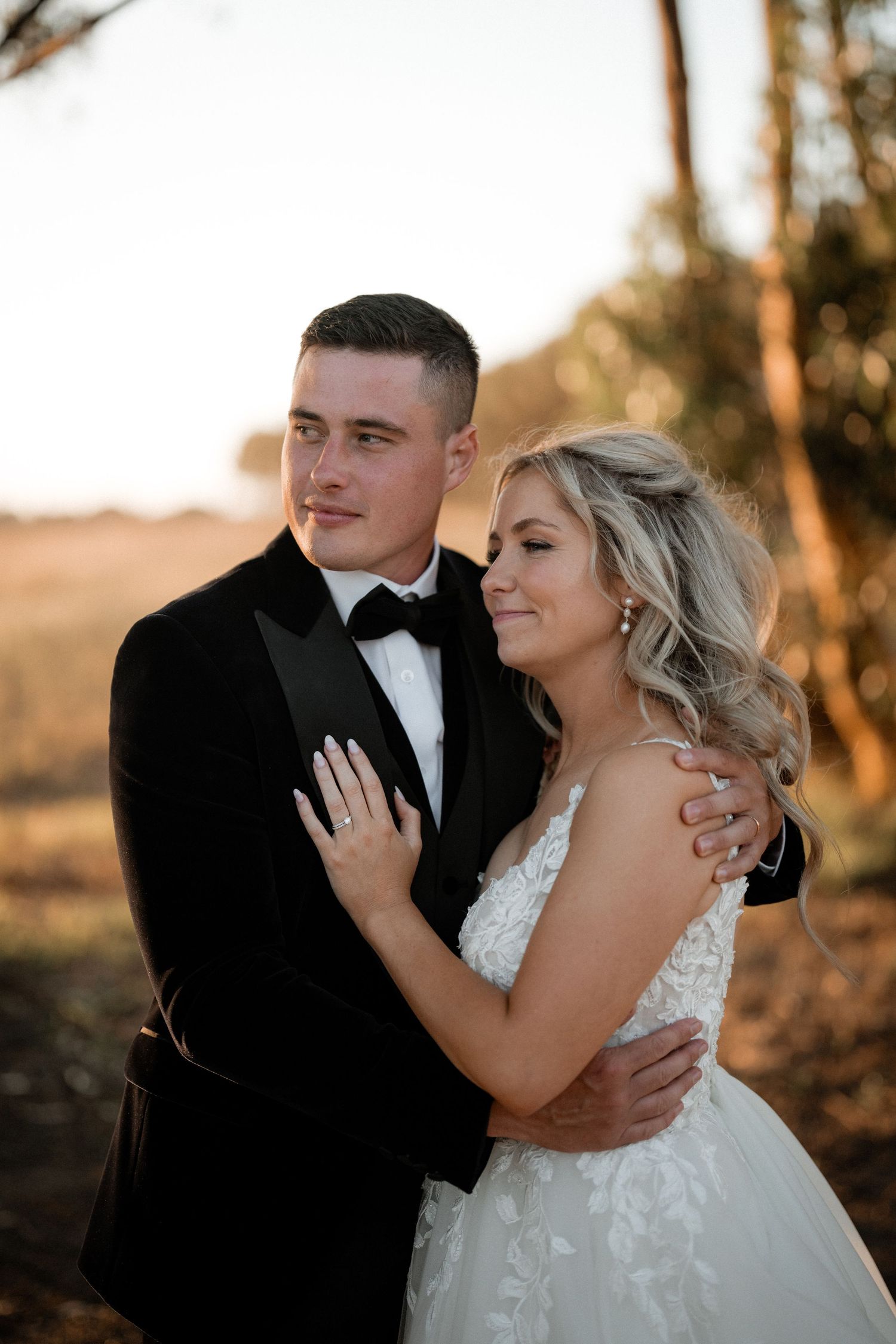 A couple embraces in formal wedding attire during golden hour outdoors.