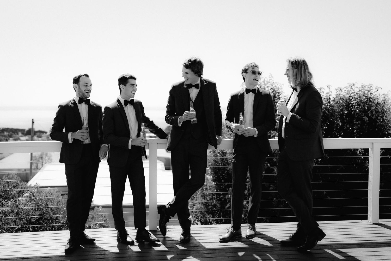 Five people dressed in formal black attire stand together chatting and drinking on a sunny rooftop terrace.