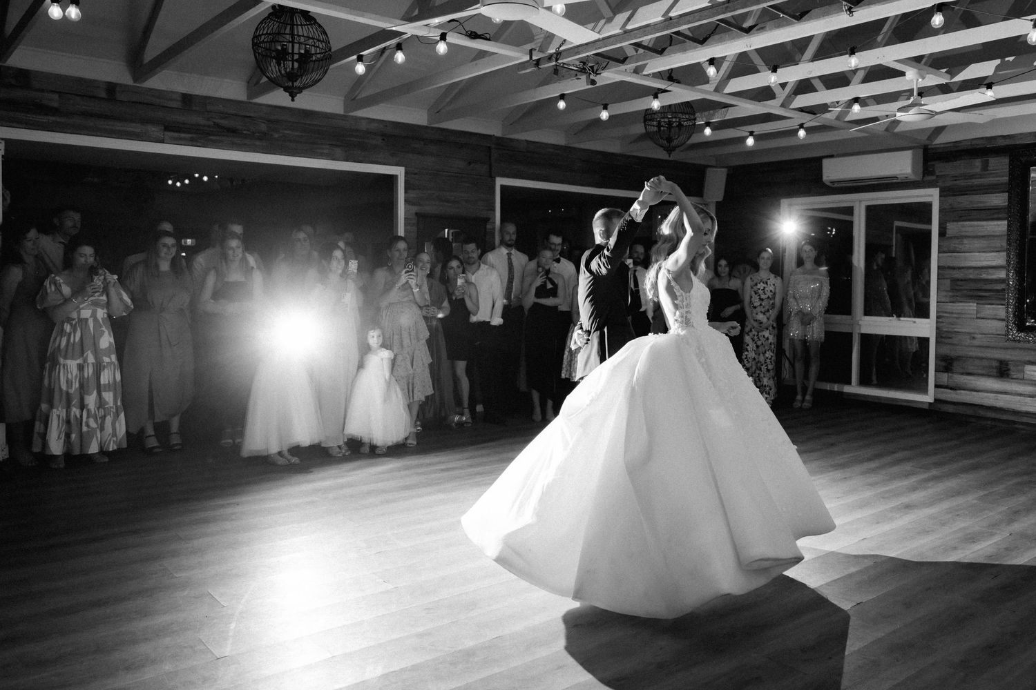 A black and white photograph shows a couple sharing their first dance at a wedding reception with string lights overhead.