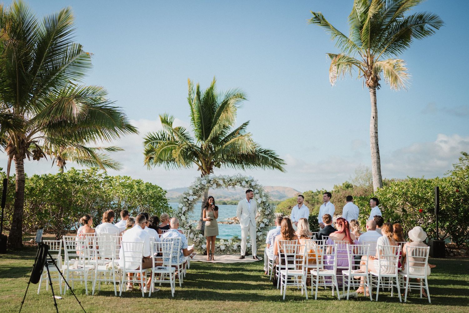 Outdoor beach wedding ceremony setup with white chairs and floral arch framed by palm trees.