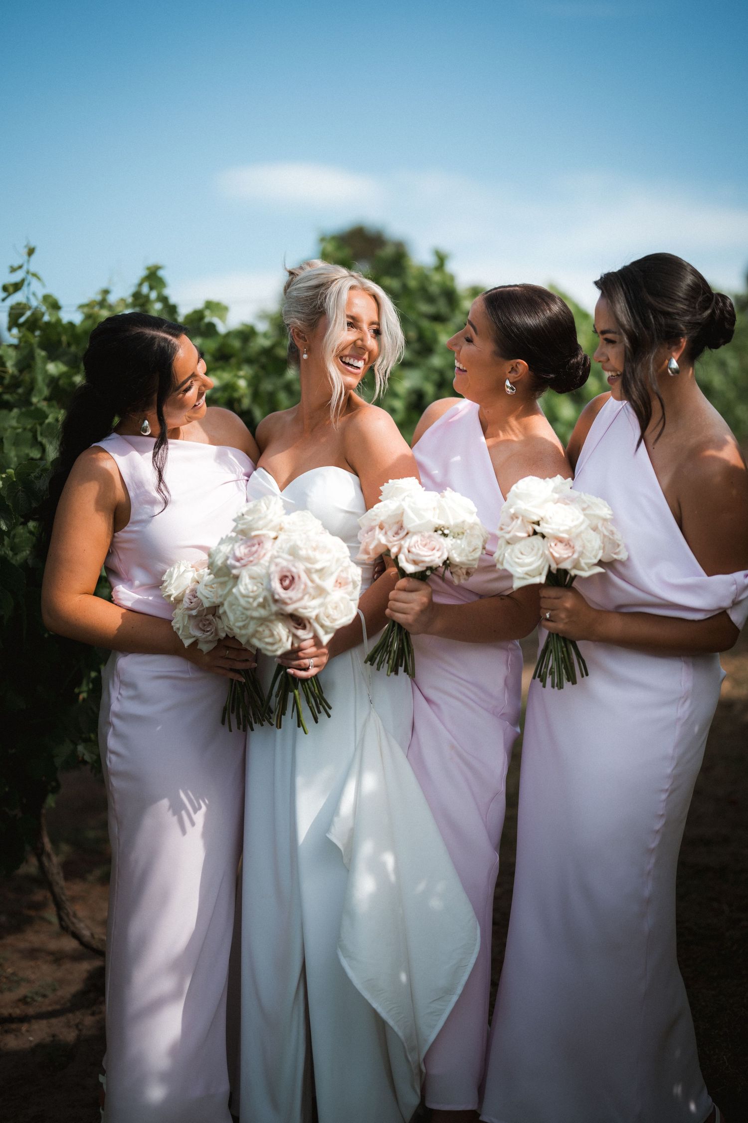 Bridal party in white dresses share a joyful moment while holding matching floral bouquets in vineyard setting.