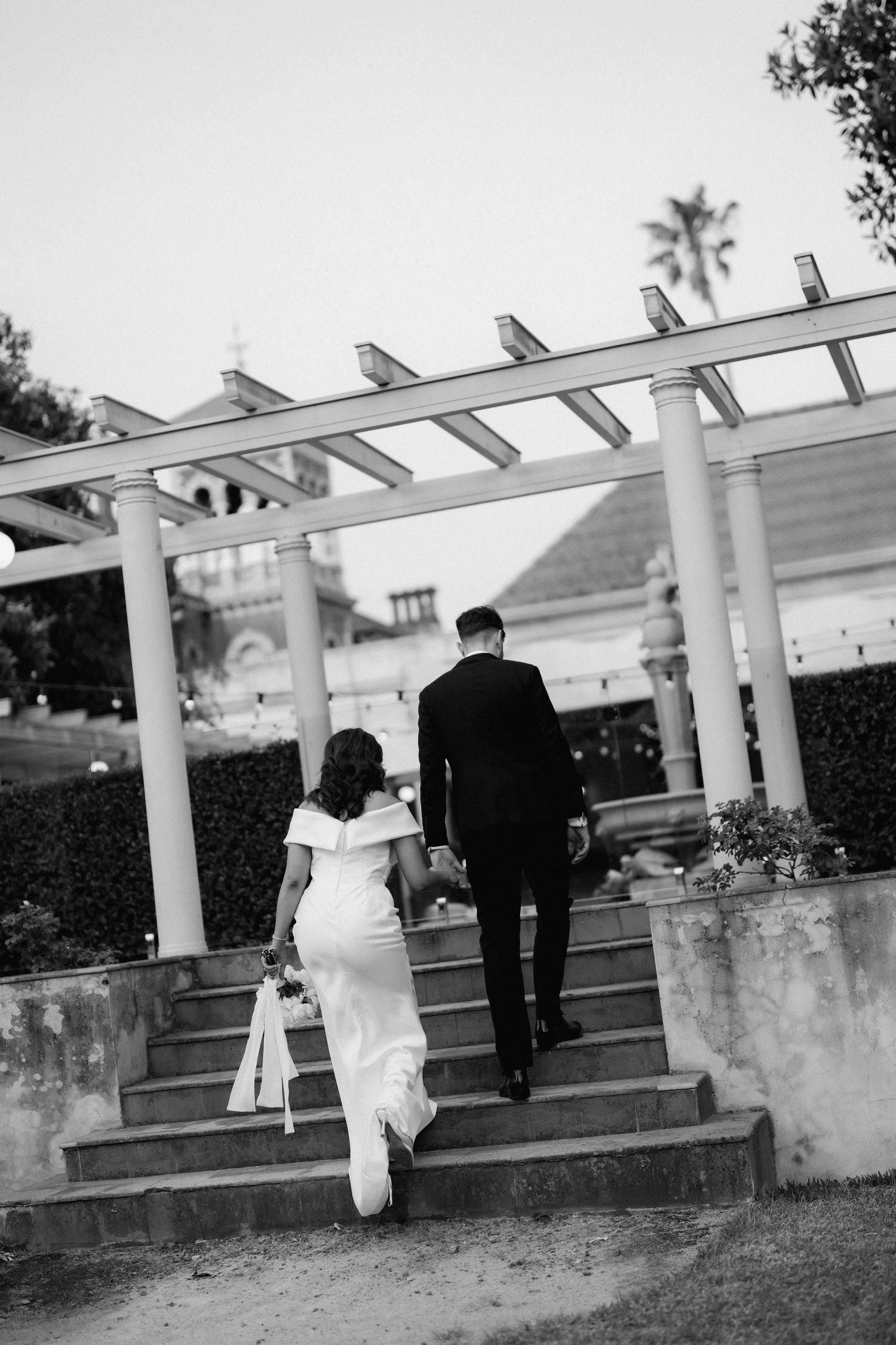 A couple in wedding attire ascending outdoor stone steps under a white pergola in black and white photography.