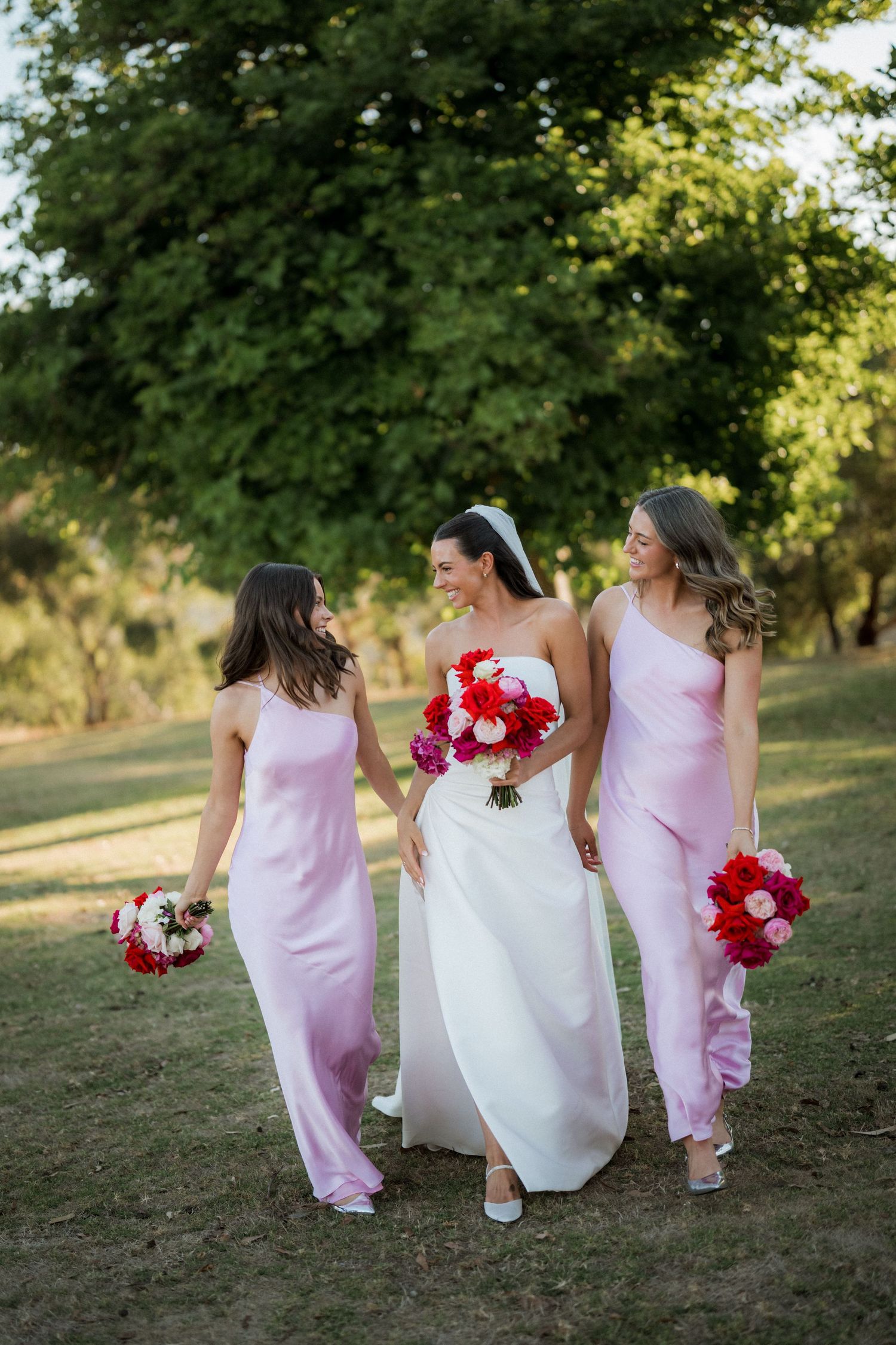 Wedding party walking together on grass with bridesmaids in pink dresses and bride in white holding red bouquets.