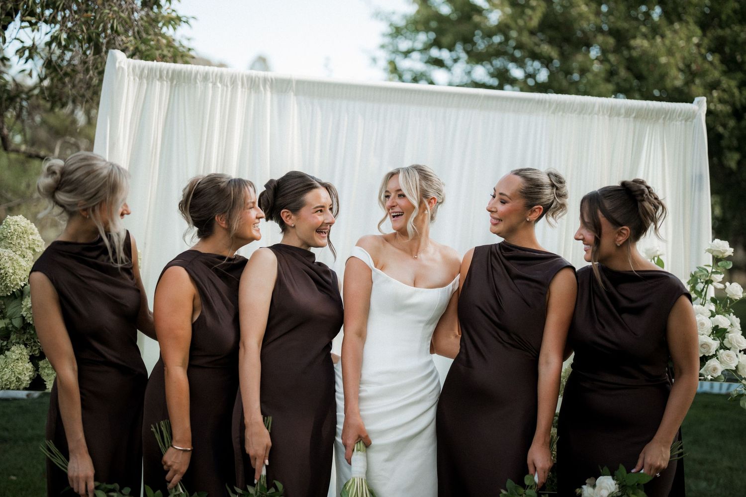 Bridesmaids in elegant black dresses stand together with bride in white against outdoor backdrop with white draping.