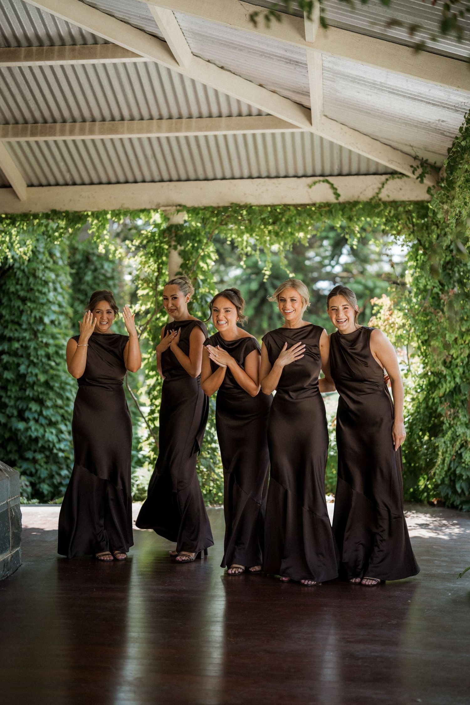 Bridesmaids in matching black dresses pose playfully together under covered outdoor area.