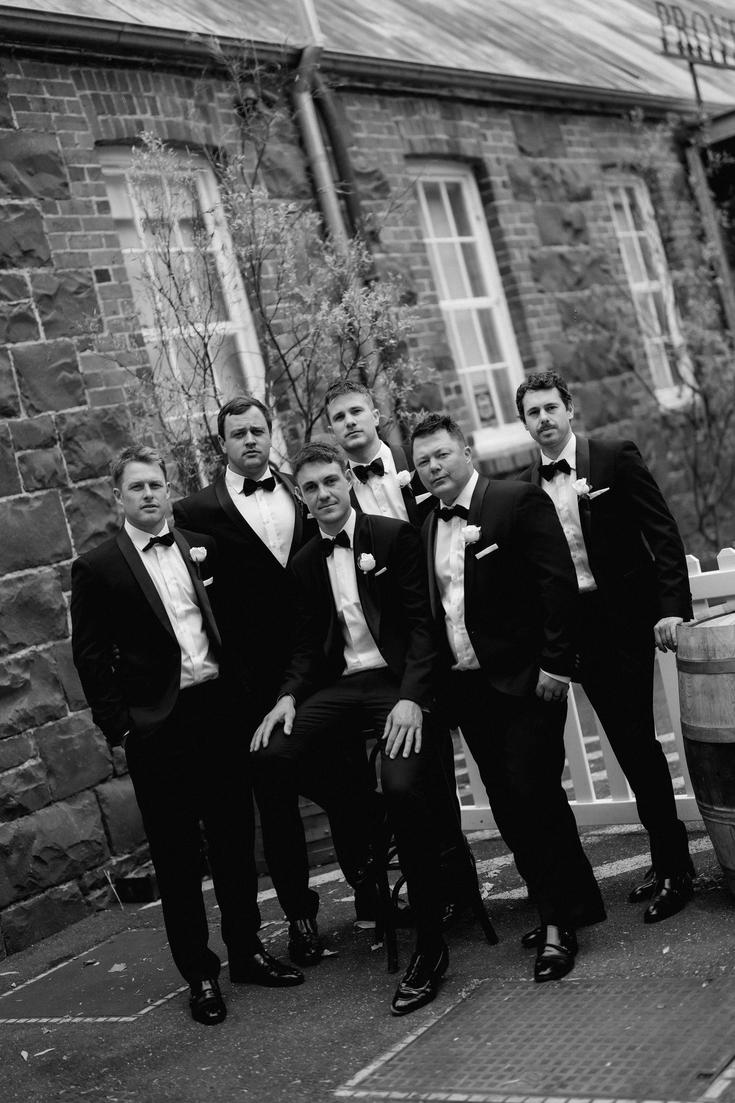 Groomsmen in black tuxedos pose casually against a rustic brick wall.