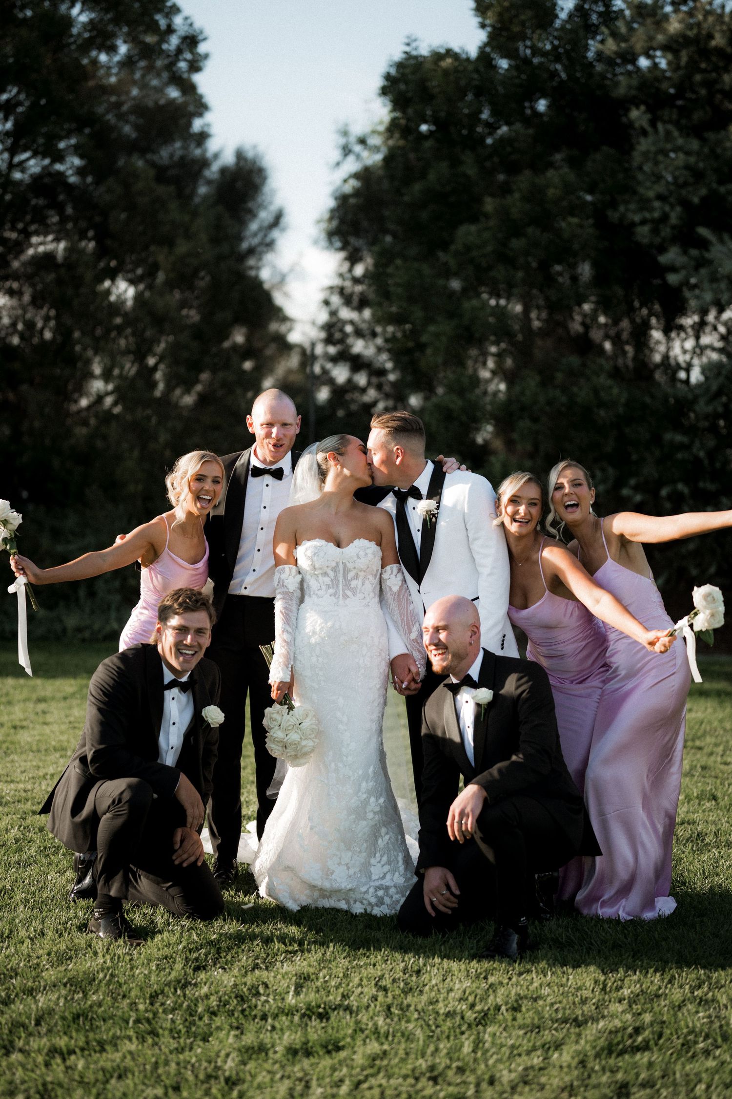 Wedding party poses playfully on grass lawn with bridesmaids in lavender dresses and groomsmen in formal attire.