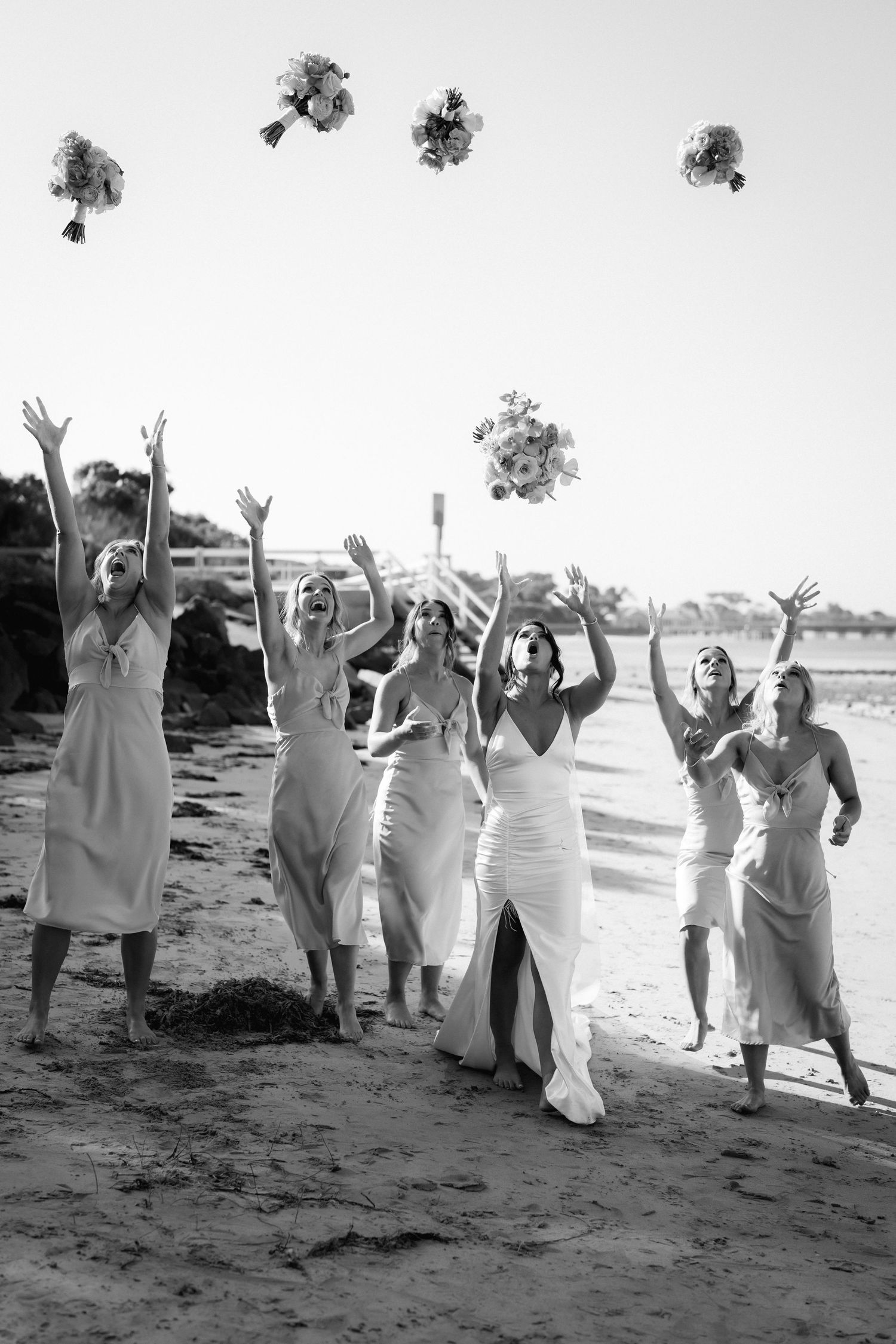 Wedding party in white attire jumps and poses joyfully on a beach in black and white photography sequence.