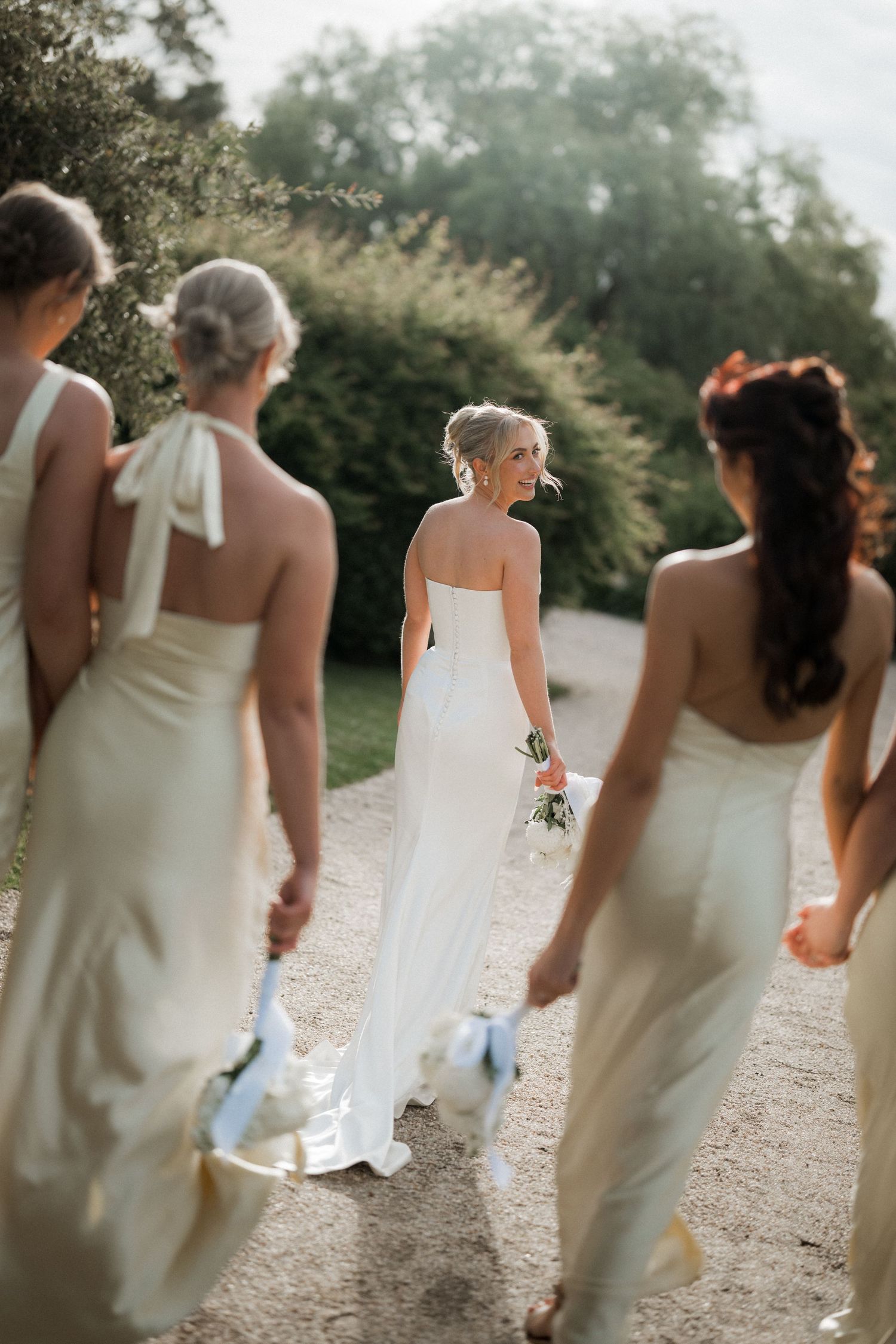 Wedding party in cream-colored dresses walking along an outdoor path in soft lighting.