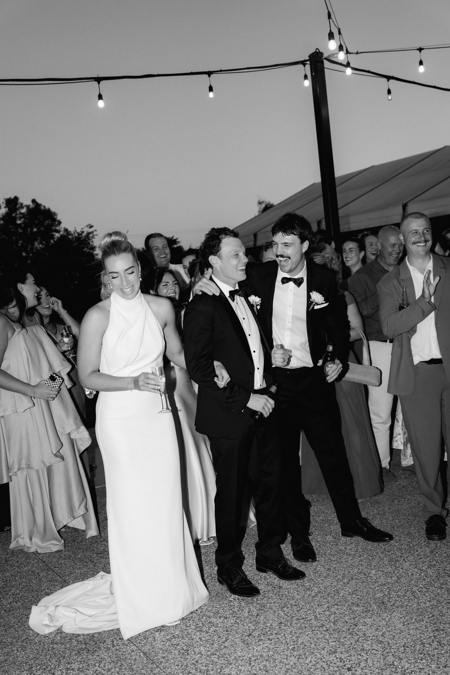 Black and white reception photo showing guests dancing under string lights at an outdoor venue.