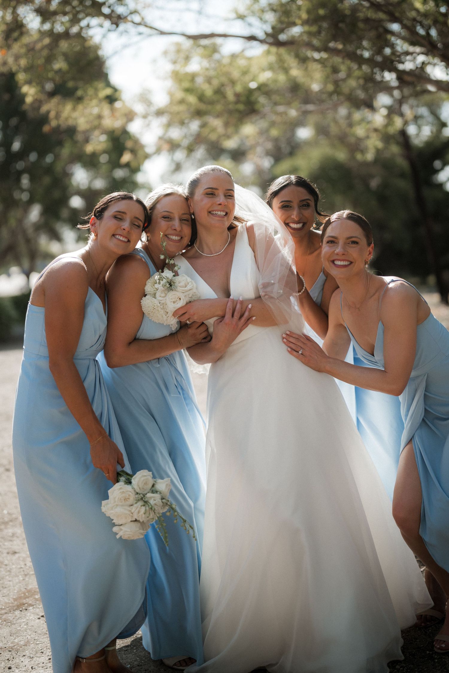 Wedding party in light blue dresses share a candid laughing moment outdoors.