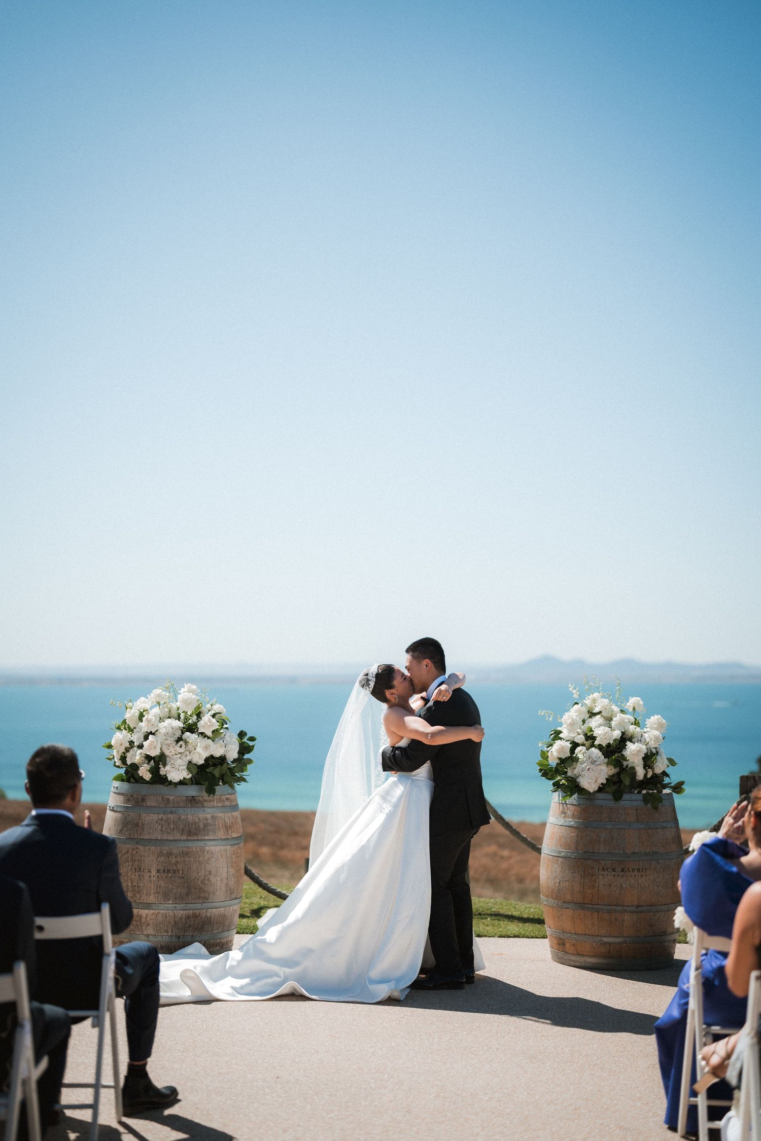 A seaside wedding ceremony features wine barrels with white floral arrangements against a blue ocean backdrop.