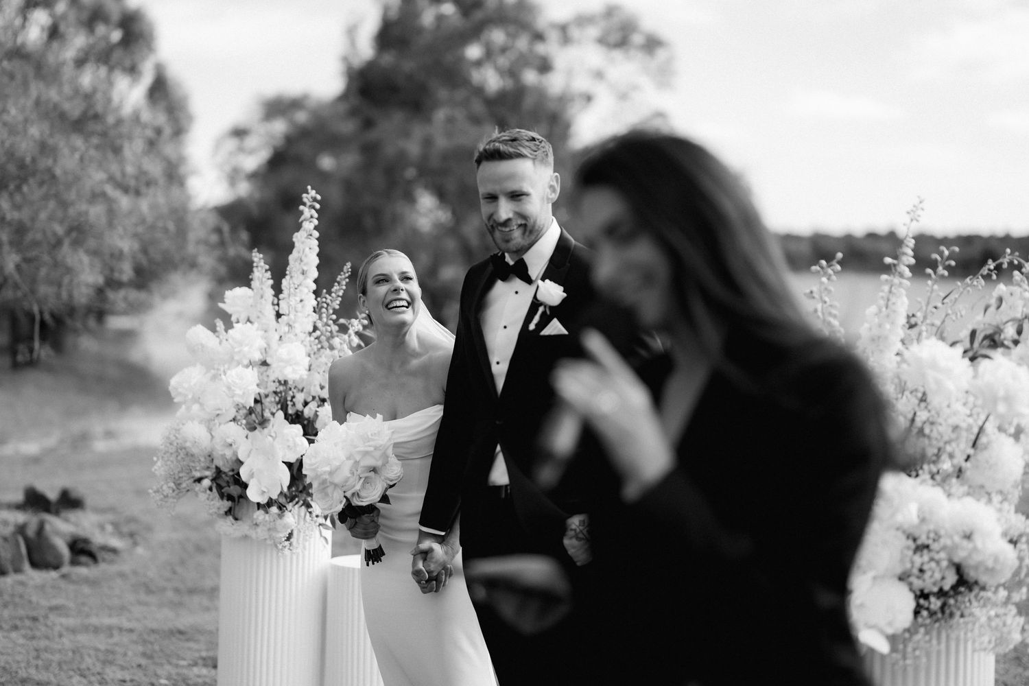 Black and white wedding photo showing an outdoor ceremony setup with elegant white floral arrangements and pedestals.