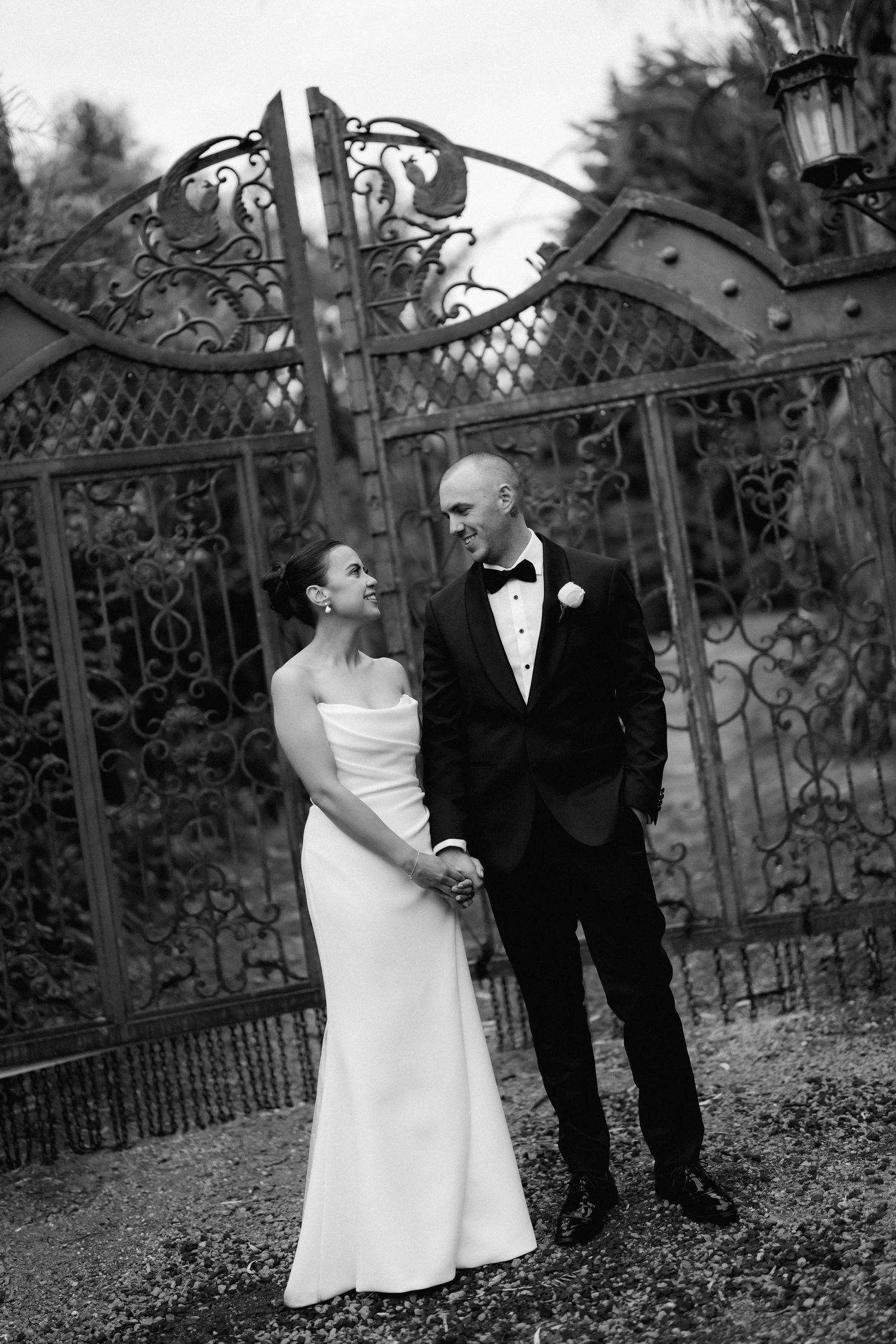 Elegant black and white wedding portrait by ornate iron gates in a garden setting.