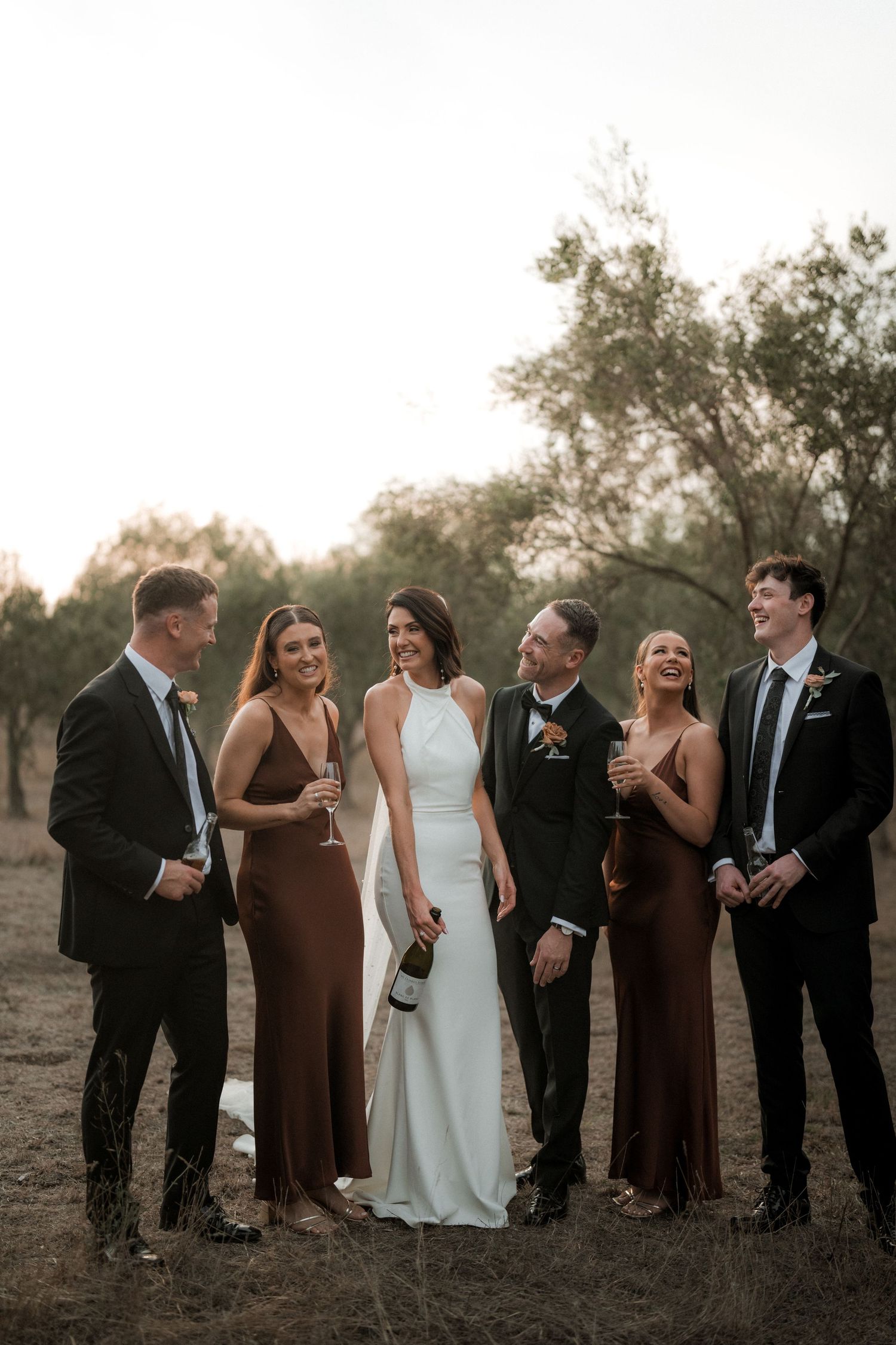 A bridal party in brown and white dresses poses together outdoors at dusk.