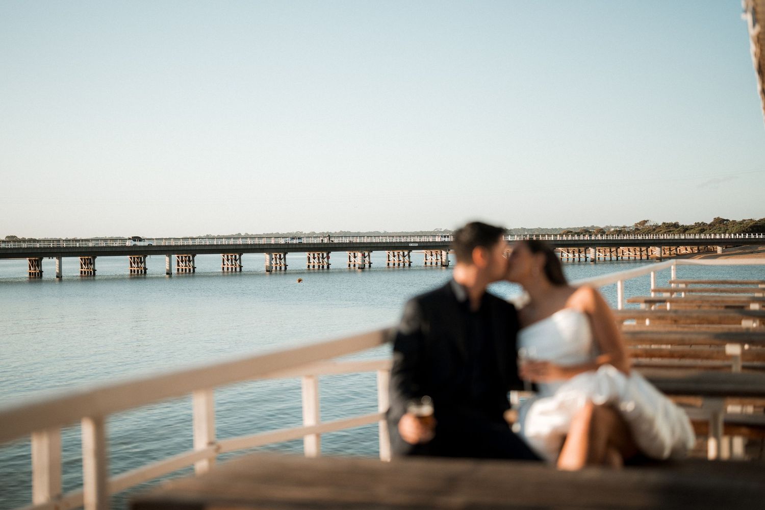 Couple sits on seaside pier bench overlooking calm blue water and distant bridge during golden hour.