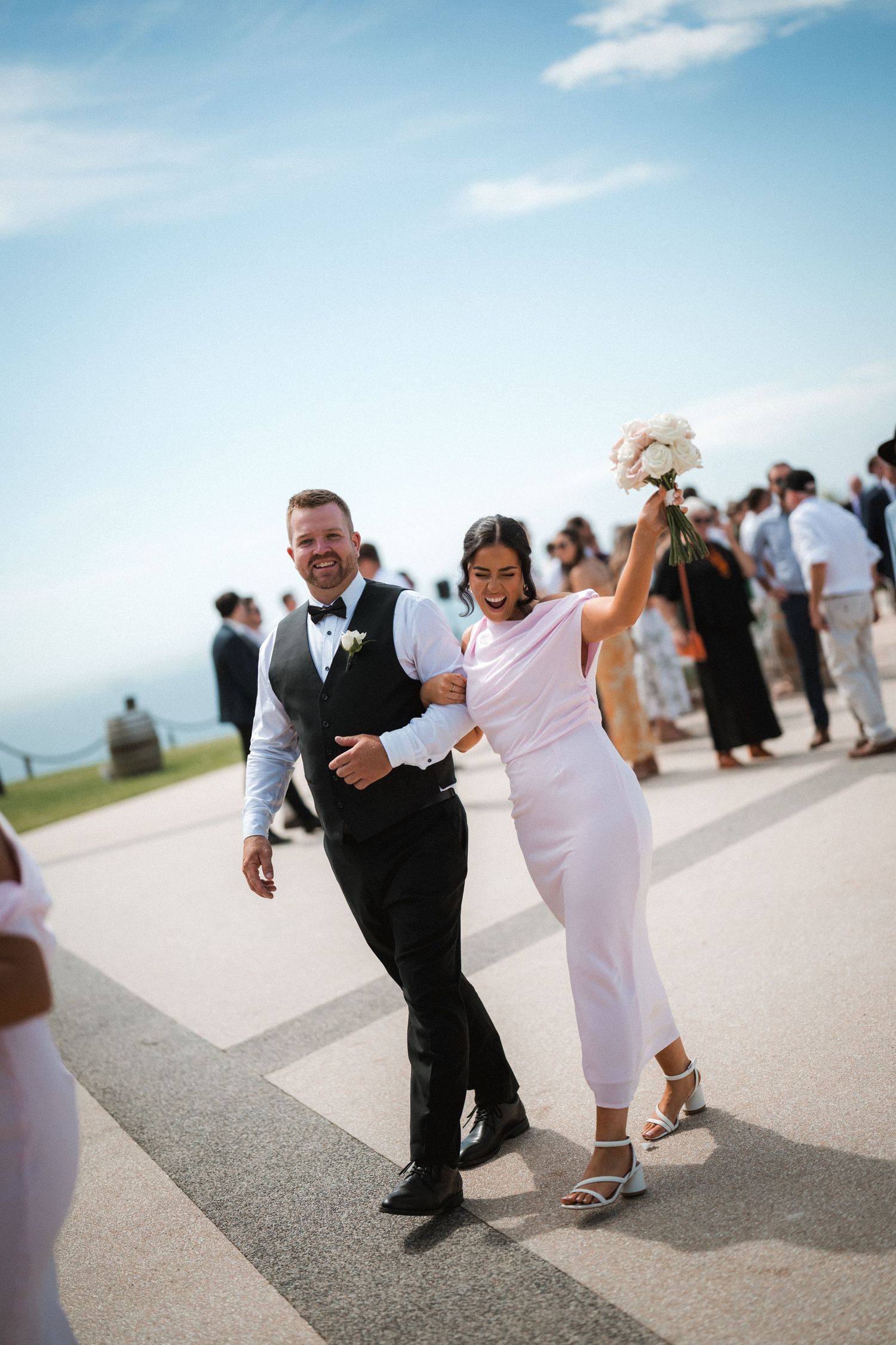 Celebratory couple dancing and walking together on a paved pathway under bright blue sky.