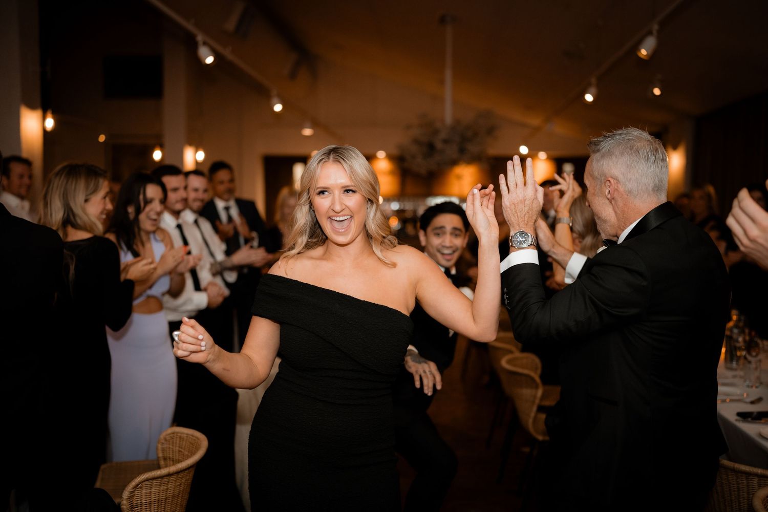 Wedding guests dancing and celebrating at an indoor reception with warm lighting.