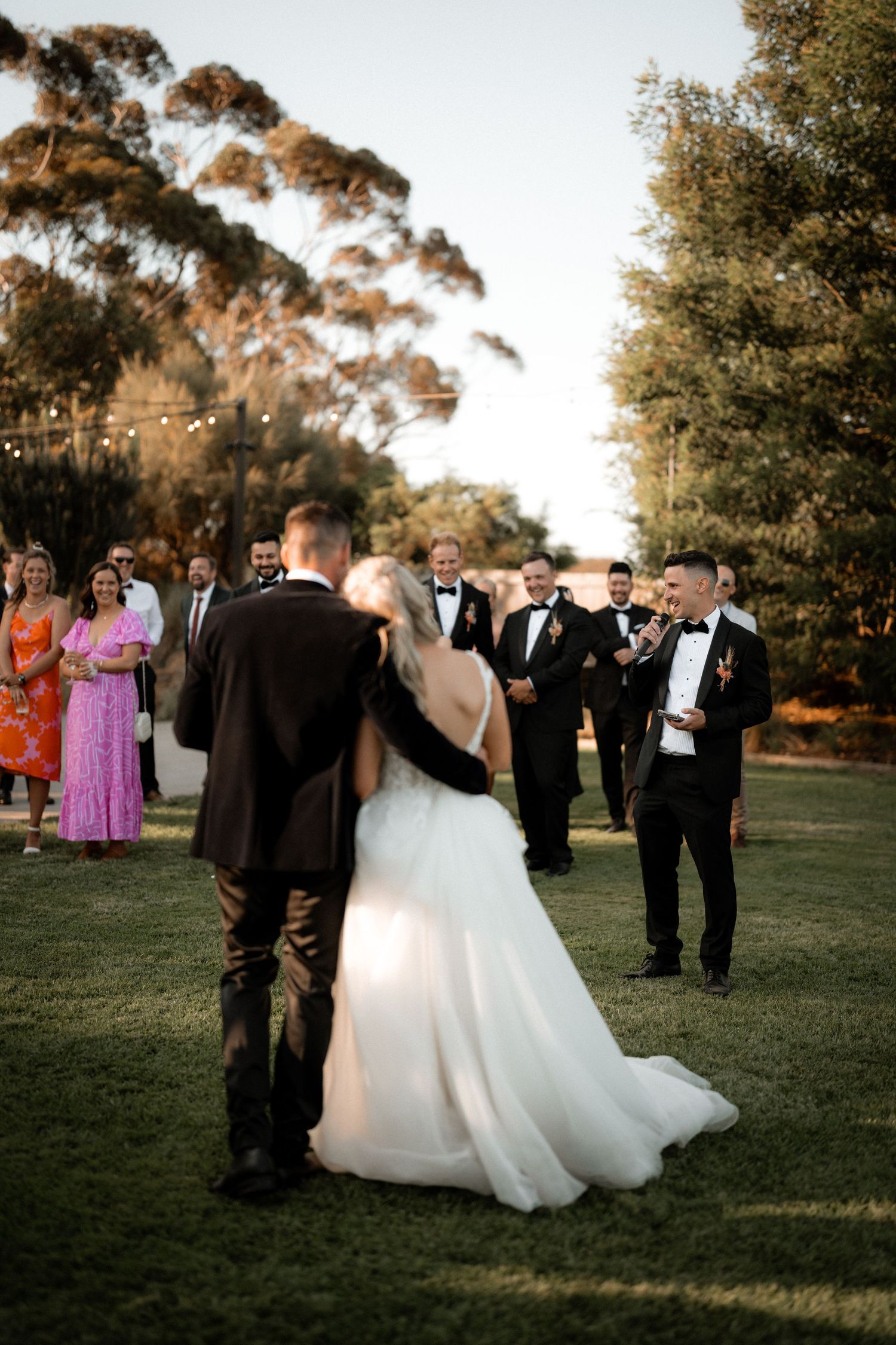Wedding party gathers on a lawn with tall eucalyptus trees during an outdoor ceremony at sunset.