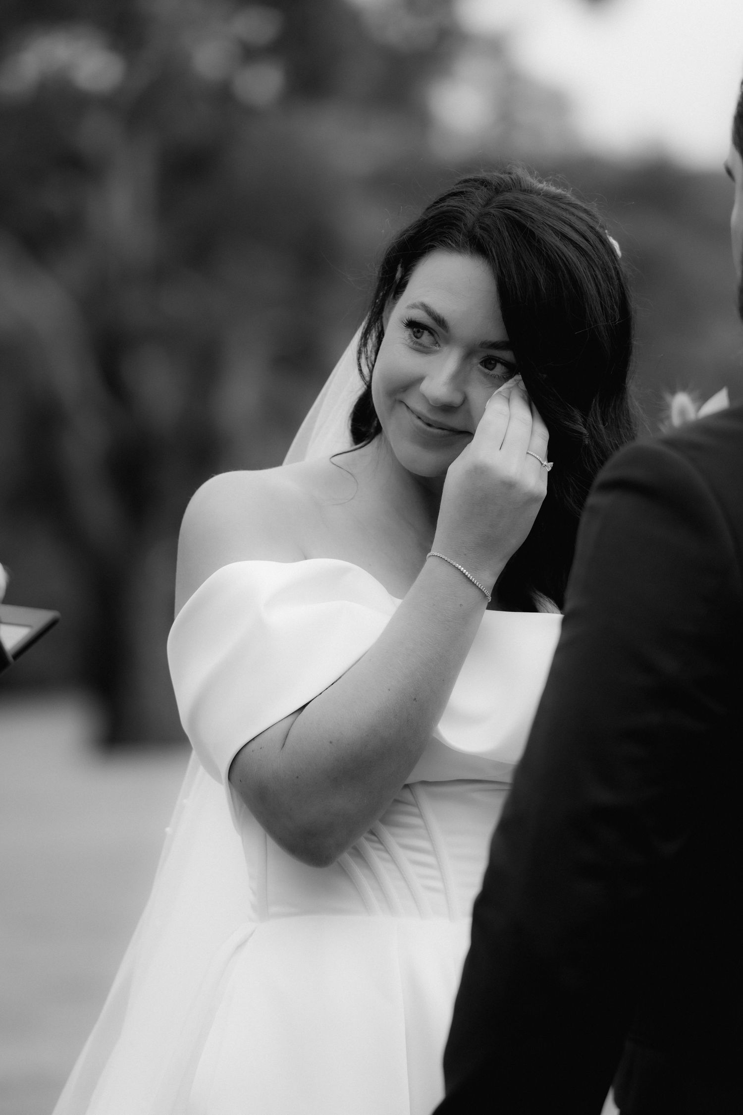 Black and white portrait of bride in off-shoulder wedding dress wiping away a tear with emotional expression.