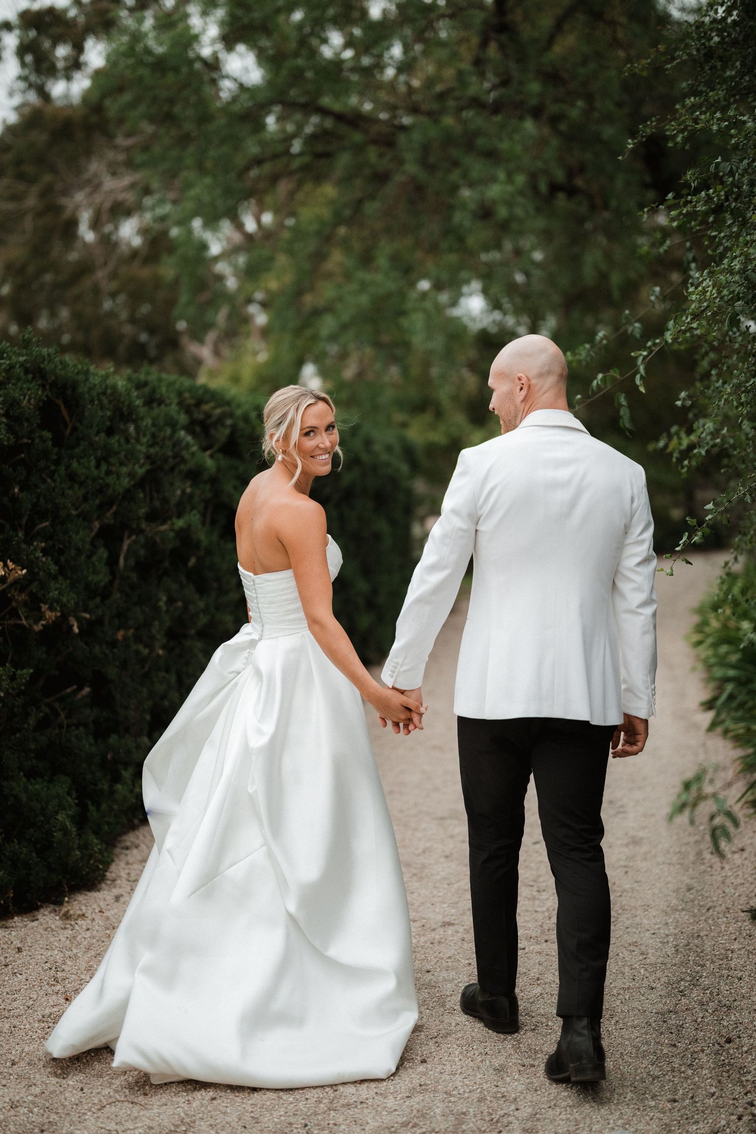 A newlywed couple holding hands while walking down a garden path with lush greenery surrounding them.