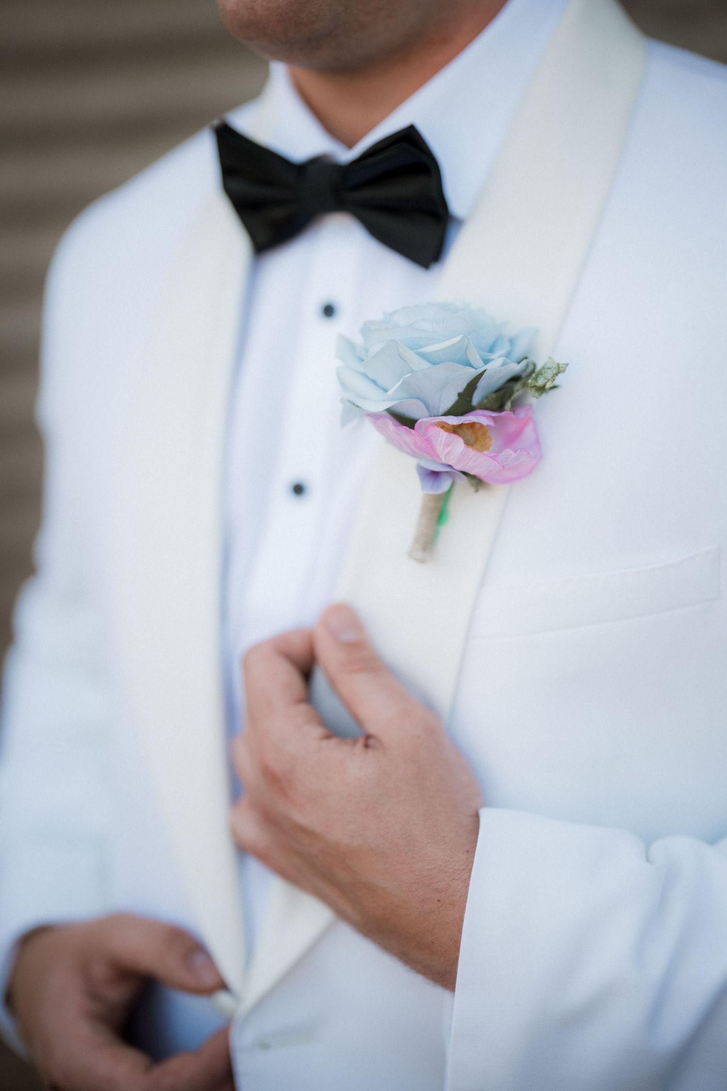 Groom in white tuxedo jacket with black bow tie adjusting light blue boutonniere on lapel.