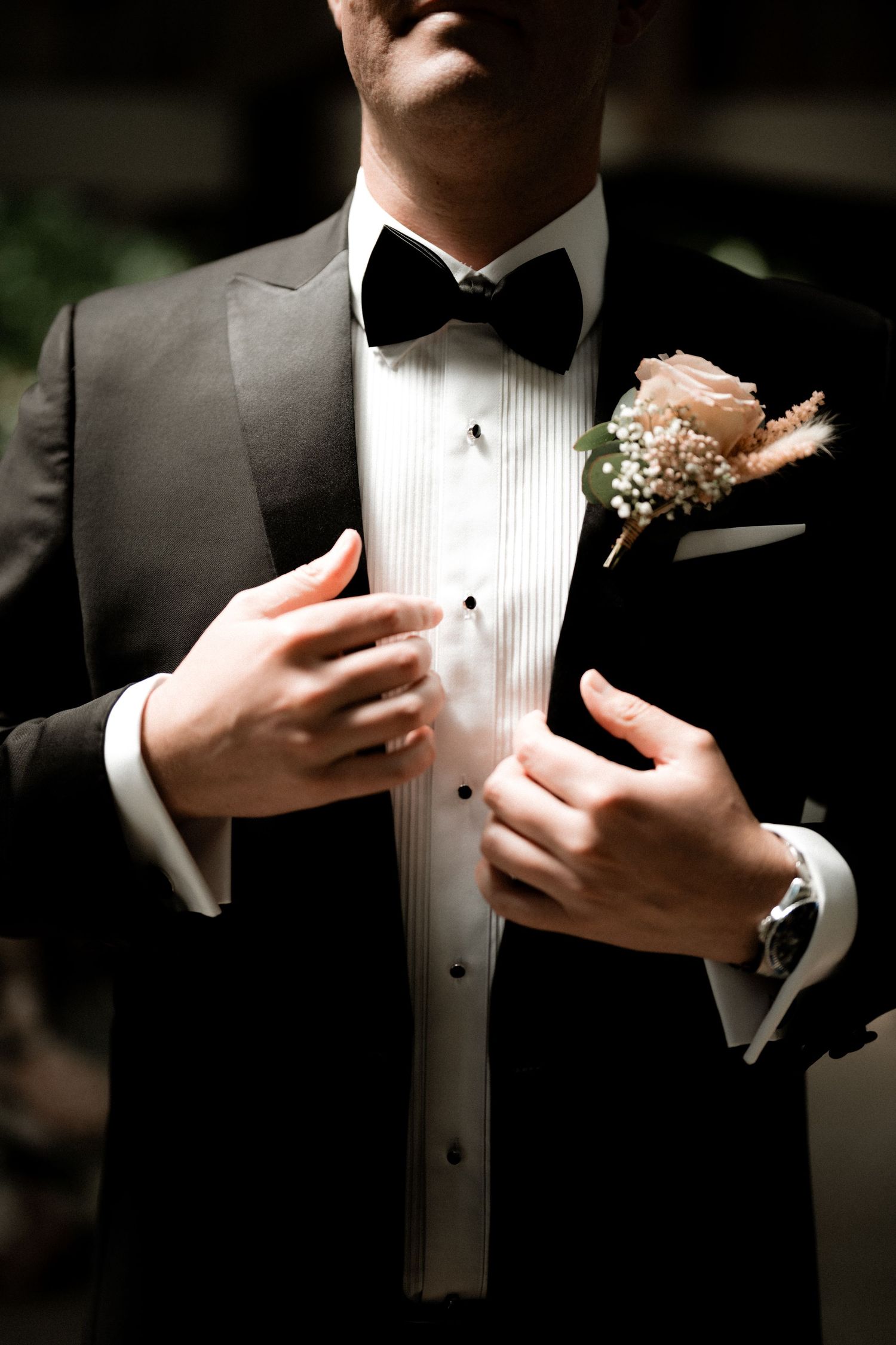 Classic groom's attire with black tuxedo, bow tie and peach boutonniere captured in dramatic lighting.