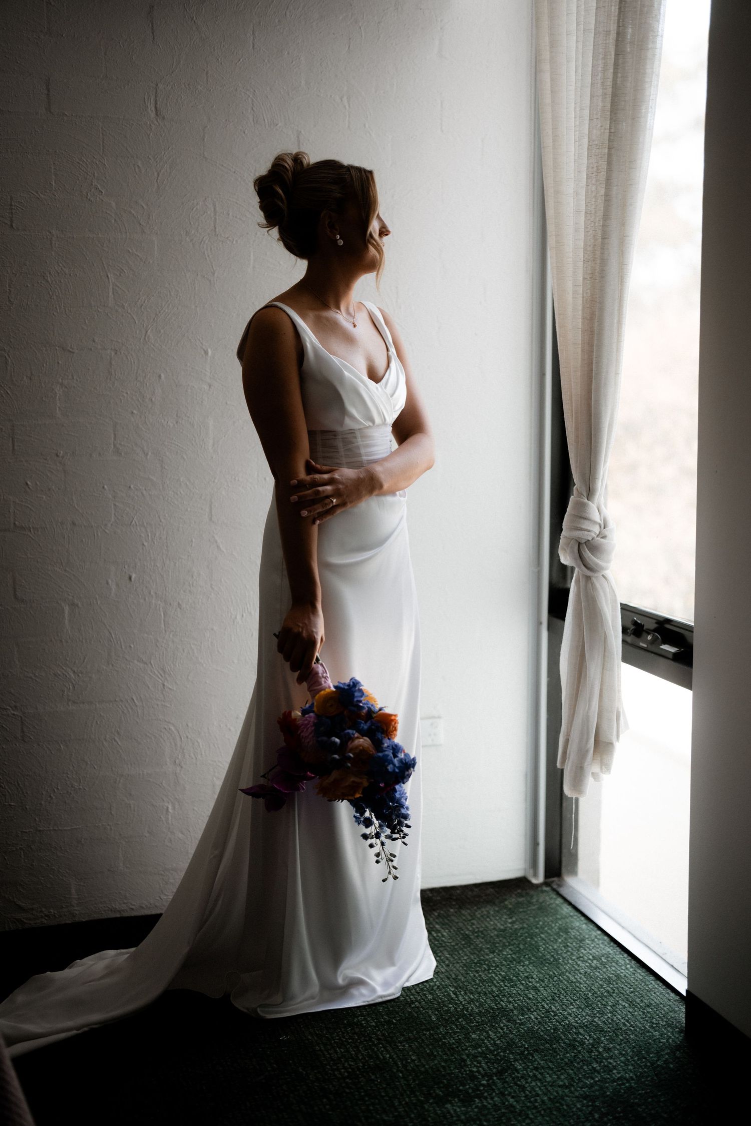 A bride in an elegant white gown stands by a window holding a vibrant blue and purple bouquet.