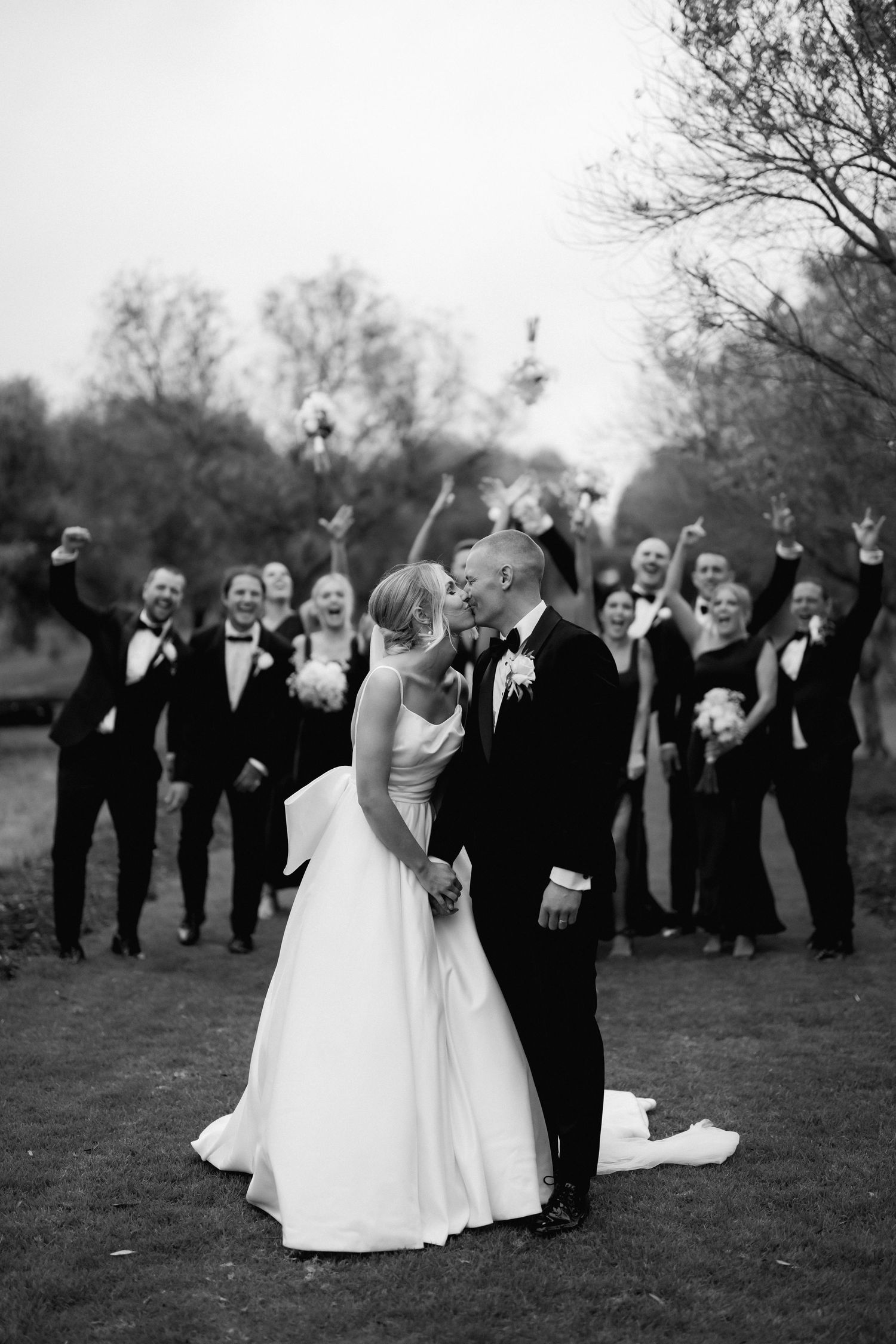 Black and white wedding photograph showing newlyweds kissing while wedding party celebrates in background.