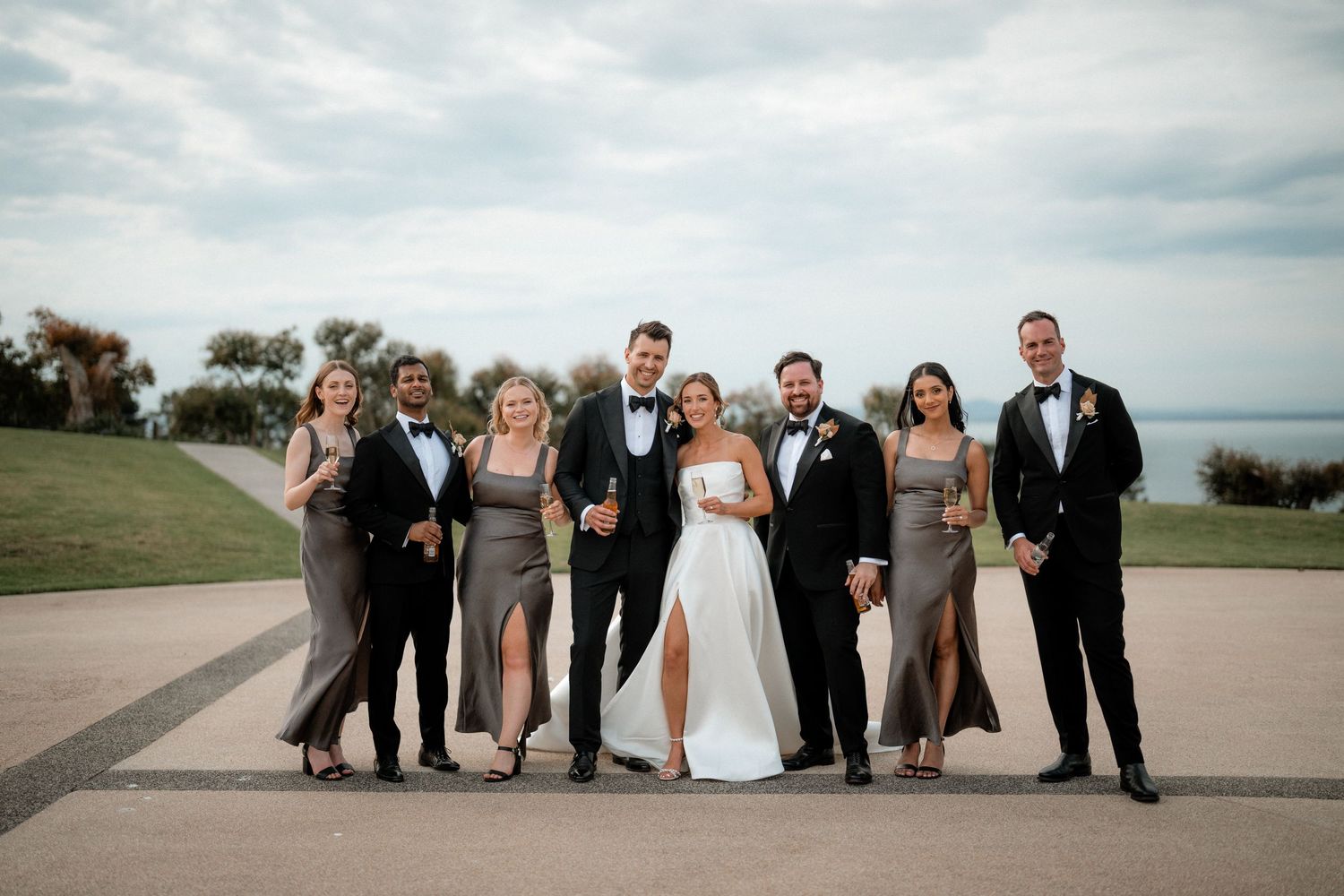 Wedding party in formal attire poses outdoors on a scenic overlook with cloudy sky and water in background.
