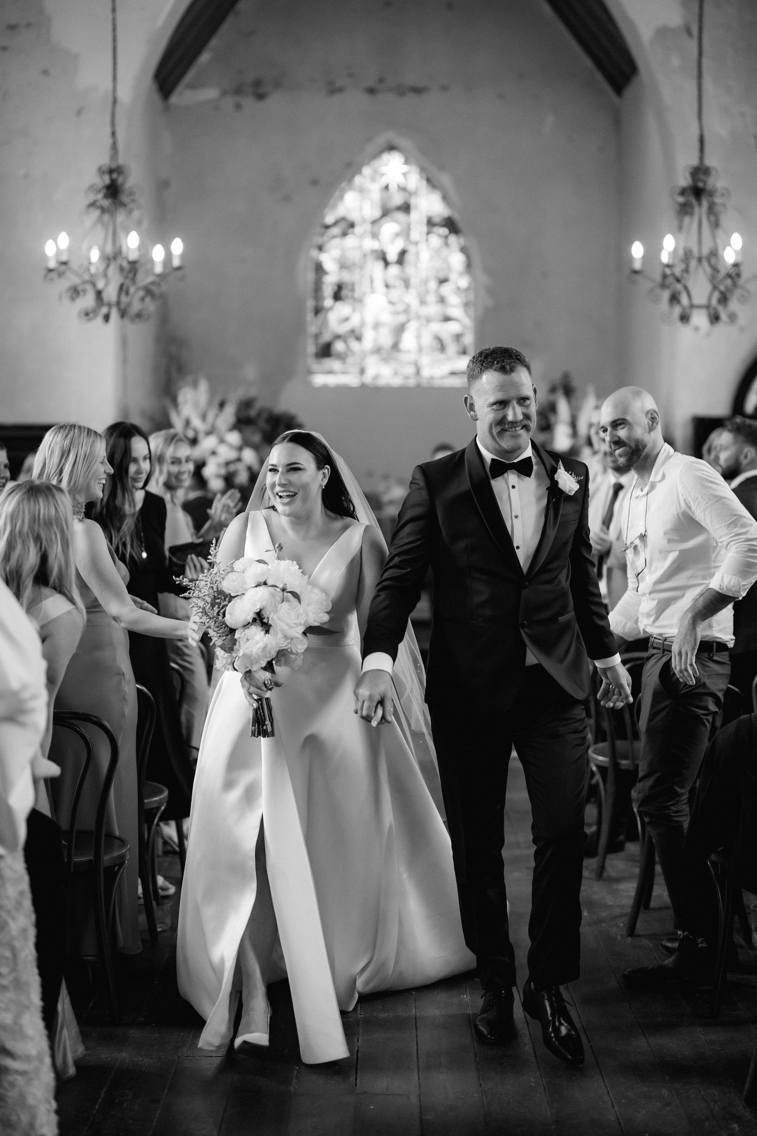 Wedding guests dance and socialize during a celebration in an elegant venue with chandeliers and vaulted ceilings.