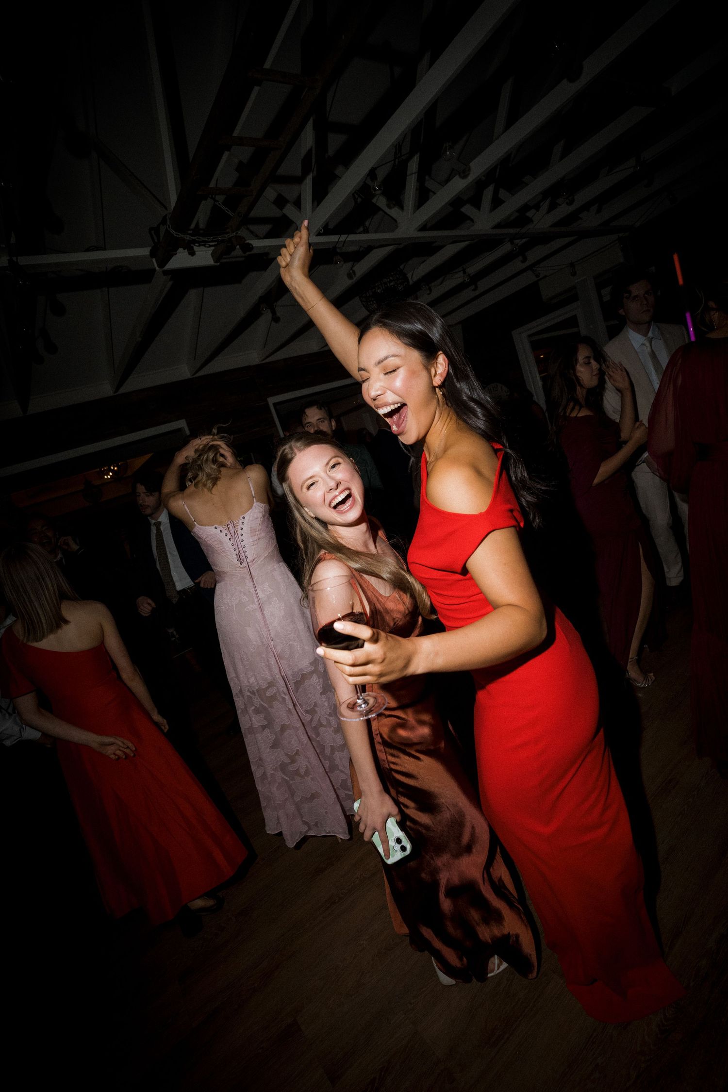 Two women in formal dresses laugh and dance together at a wedding reception.