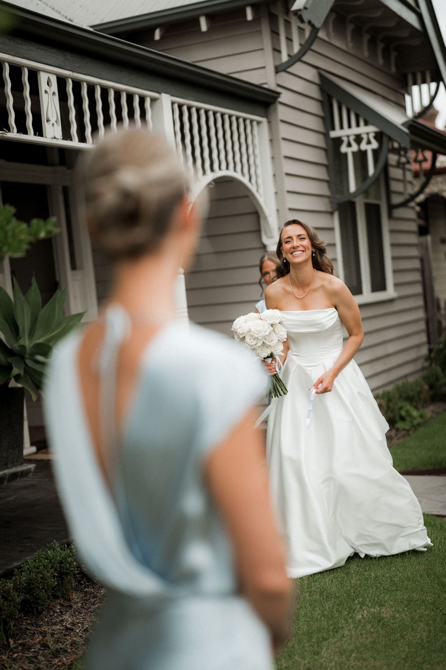 A bride in a flowing white gown poses gracefully on the lawn of a classic house with bridesmaids in blue.