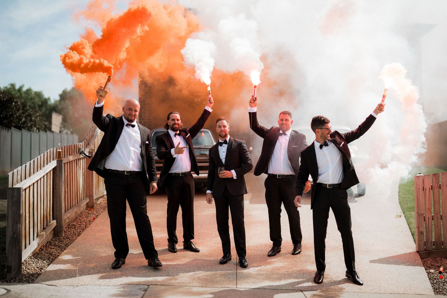 Groomsmen in formal attire posing with orange smoke bombs creating dramatic effect outdoors.
