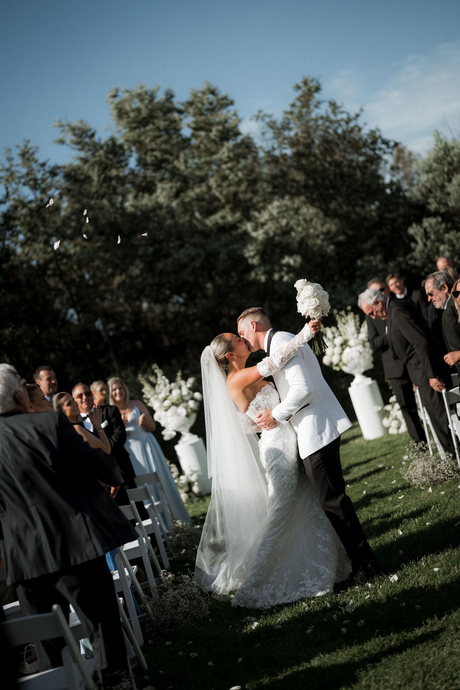 Wedding guests gather on a grassy lawn during an outdoor ceremony under blue skies and trees.