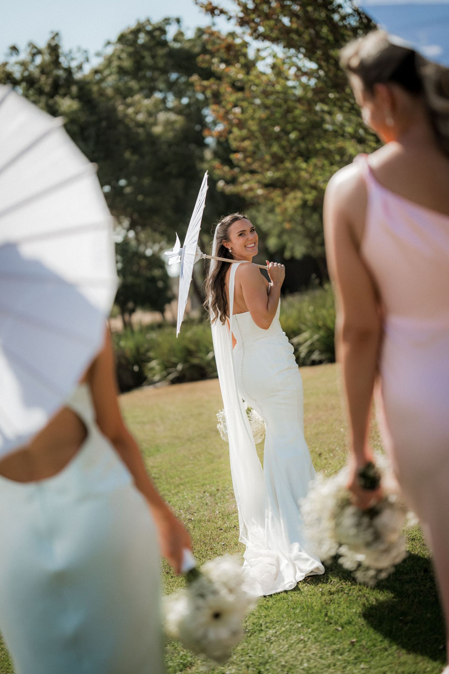 Bridesmaids in light colored dresses holding bouquets stand together on a sunny lawn during an outdoor wedding ceremony.