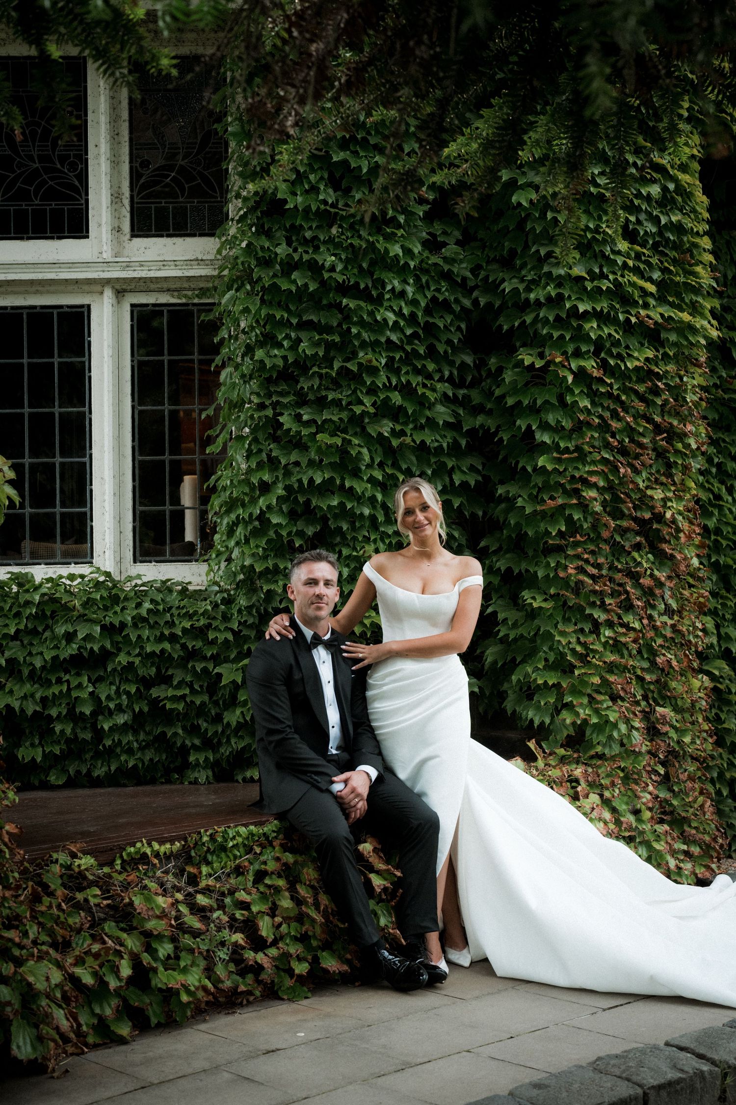 A couple in formal wedding attire poses against an ivy-covered wall with windows.