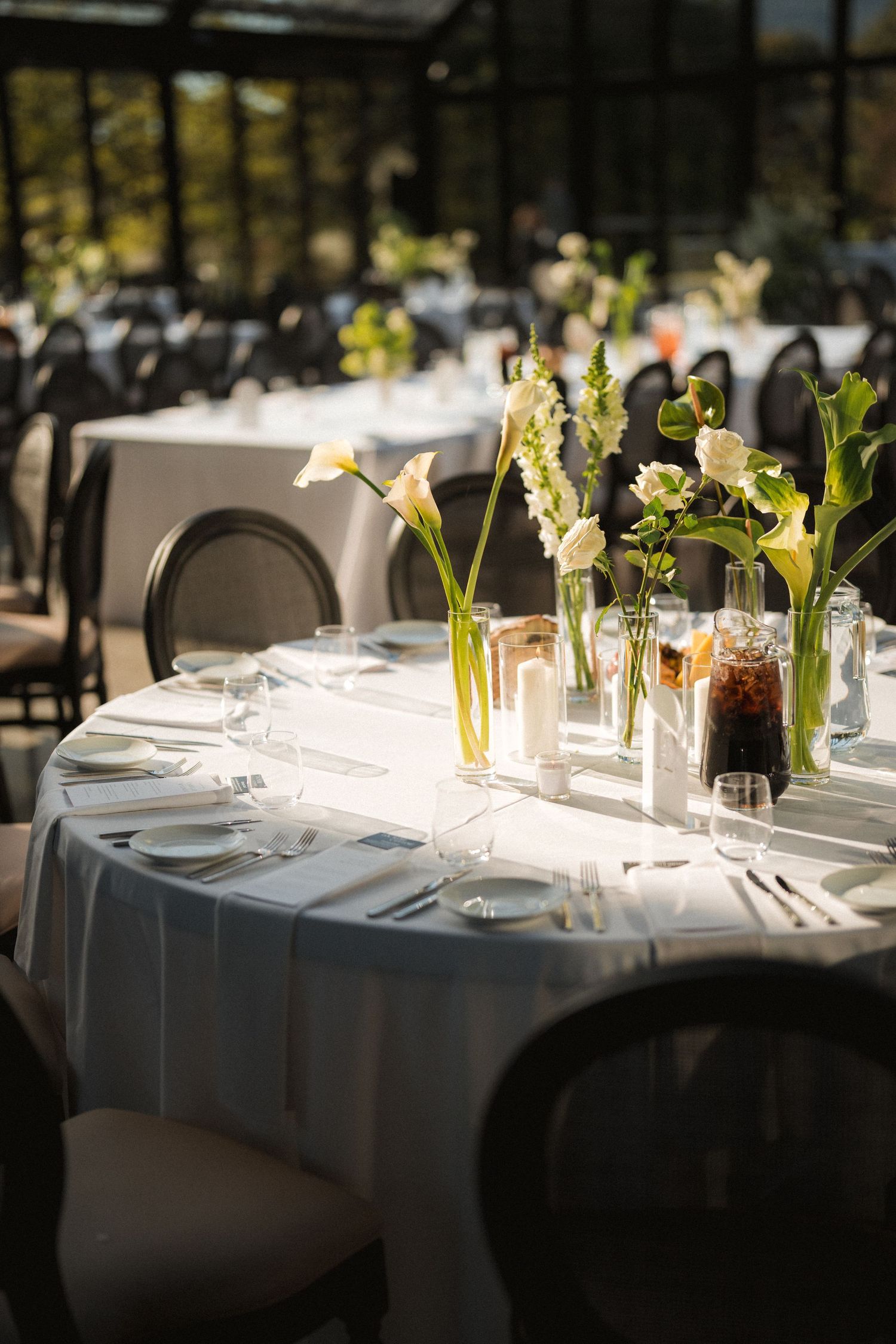Elegant round wedding reception table with white linens, tall calla lily vases, candles and place settings inside a bright greenhouse venue.