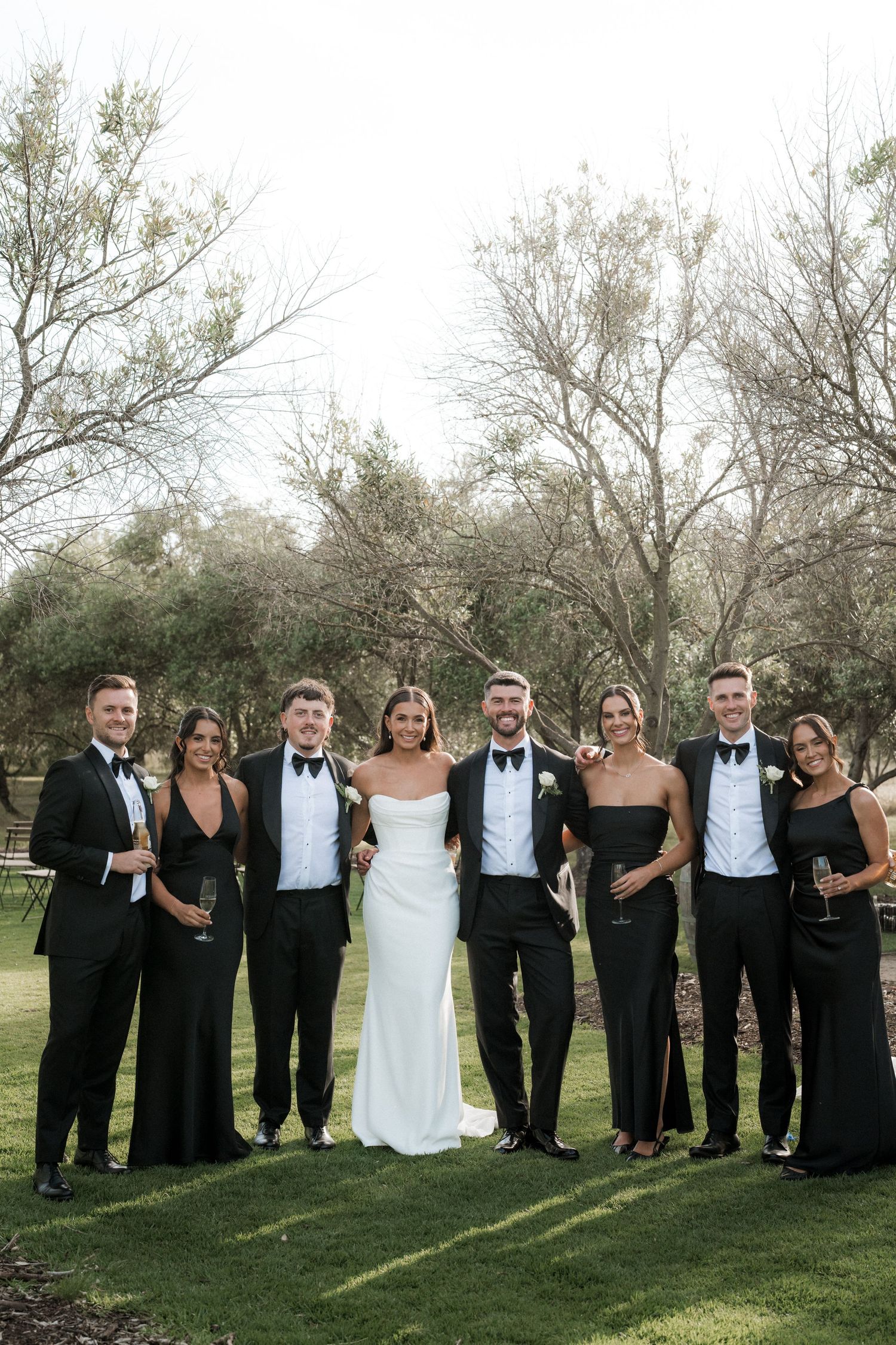 Wedding party in formal black and white attire posed together on grass.