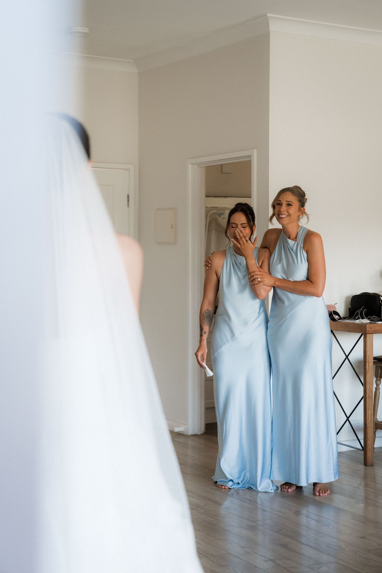 Two bridesmaids in light blue floor-length dresses react emotionally to seeing the bride in her wedding gown.