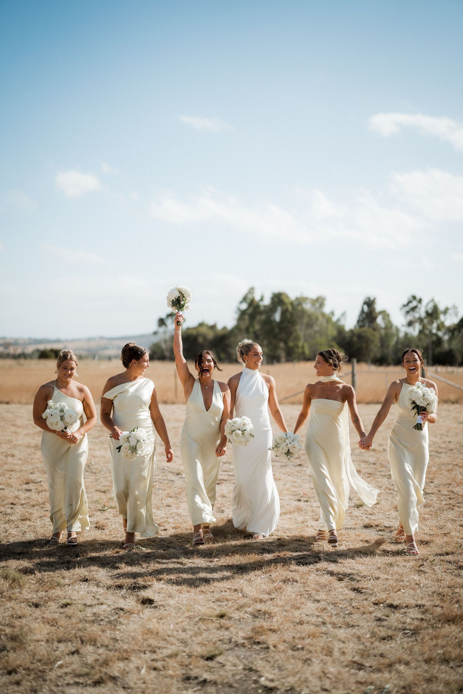 Wedding party in white dresses poses playfully on a sunlit beach landscape.