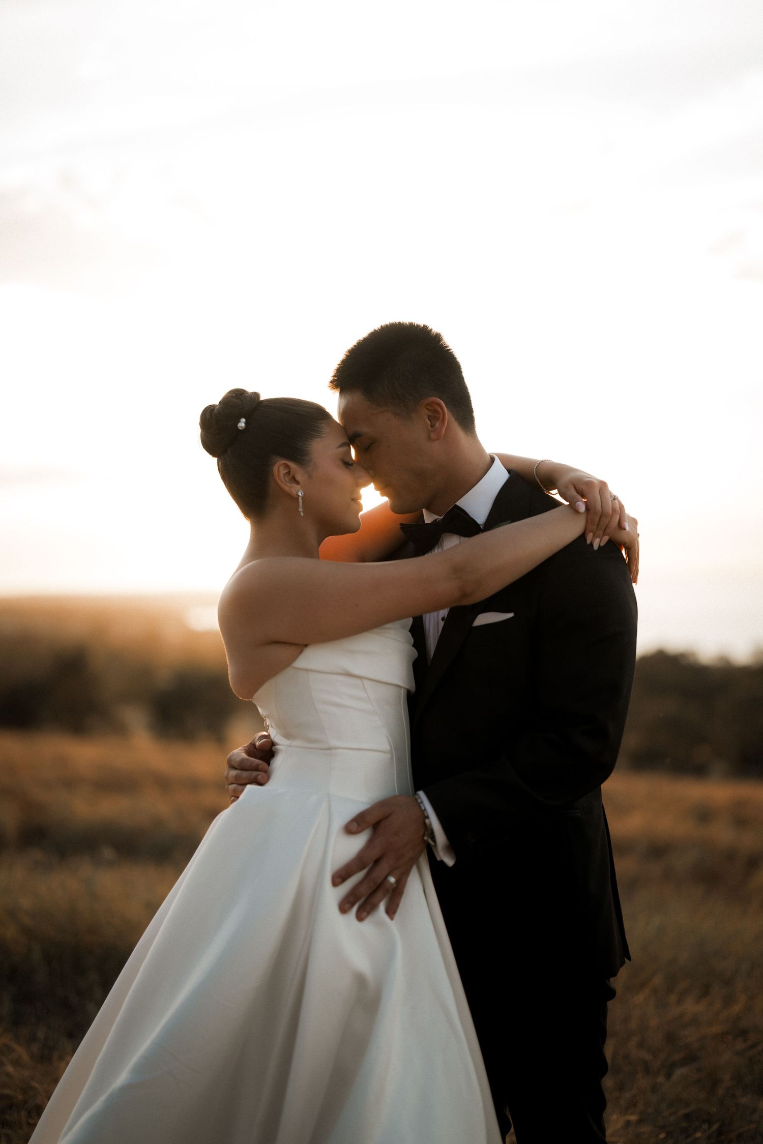A couple embraces in golden sunset light with the bride wearing an elegant white gown and groom in a dark suit.