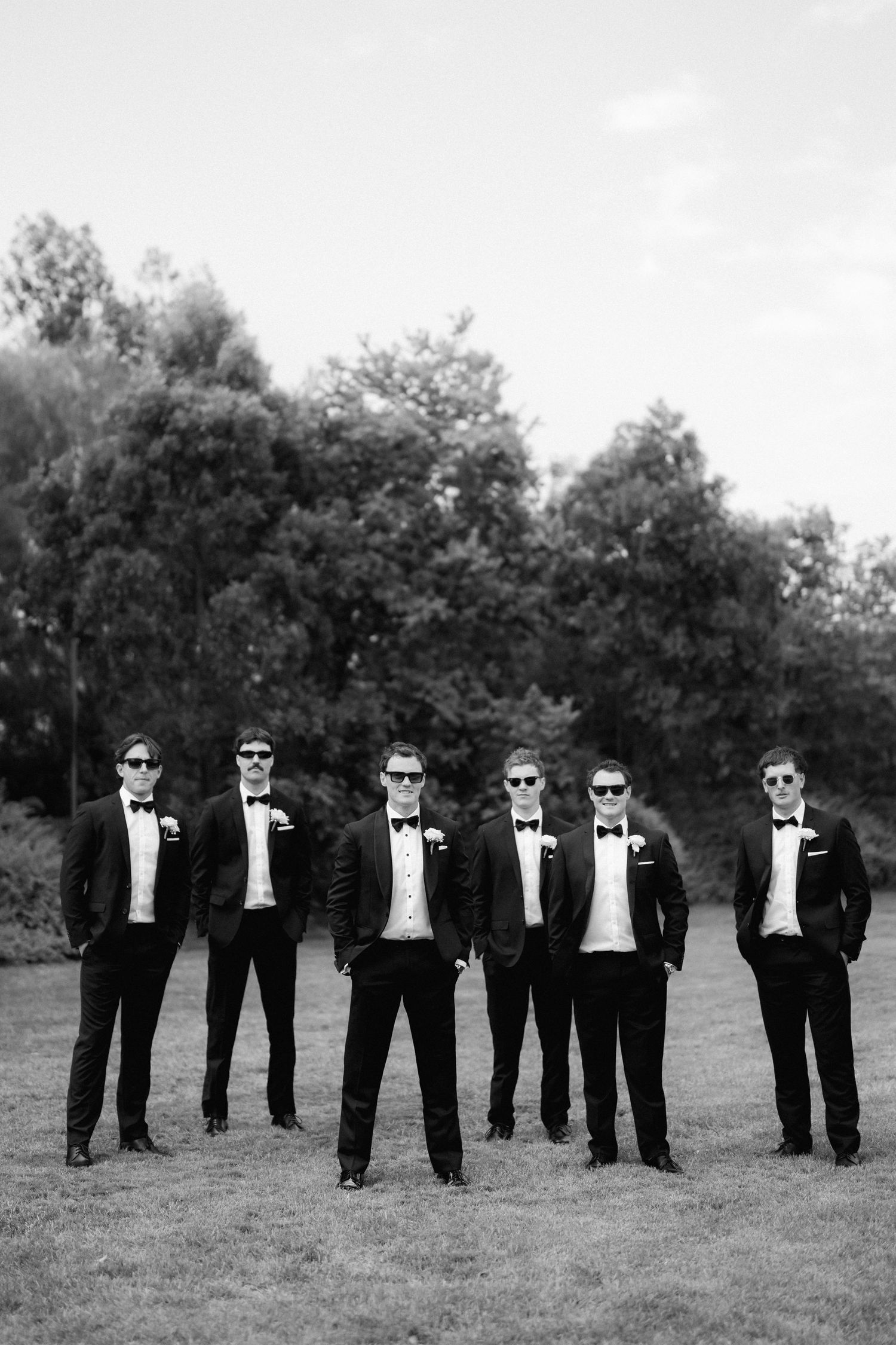 A group of groomsmen in black tuxedos and sunglasses stand in a line on grass in a stylish wedding photo.