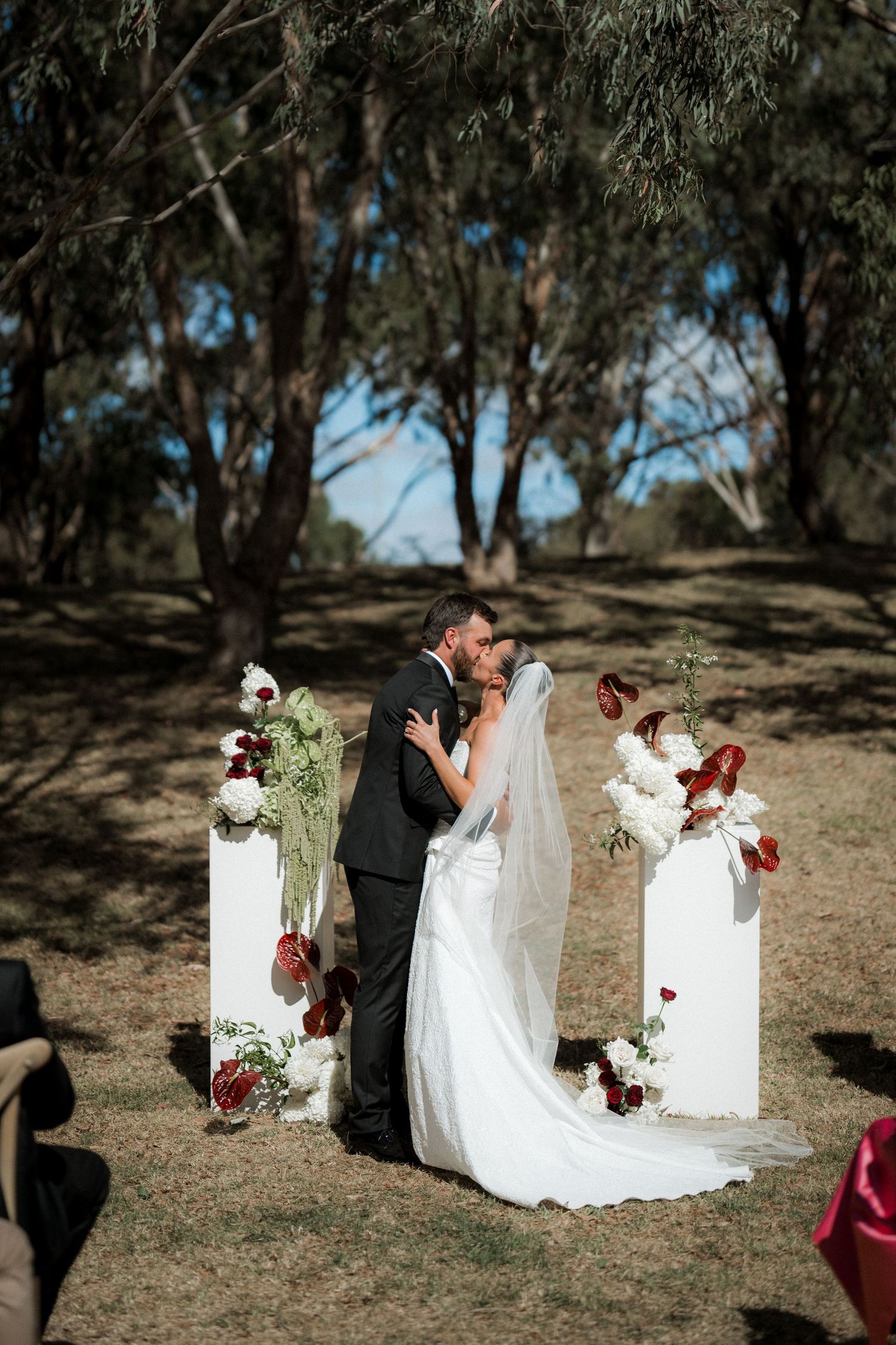 Bride and groom share first kiss at outdoor wedding ceremony with white floral pedestals and eucalyptus trees backdrop.