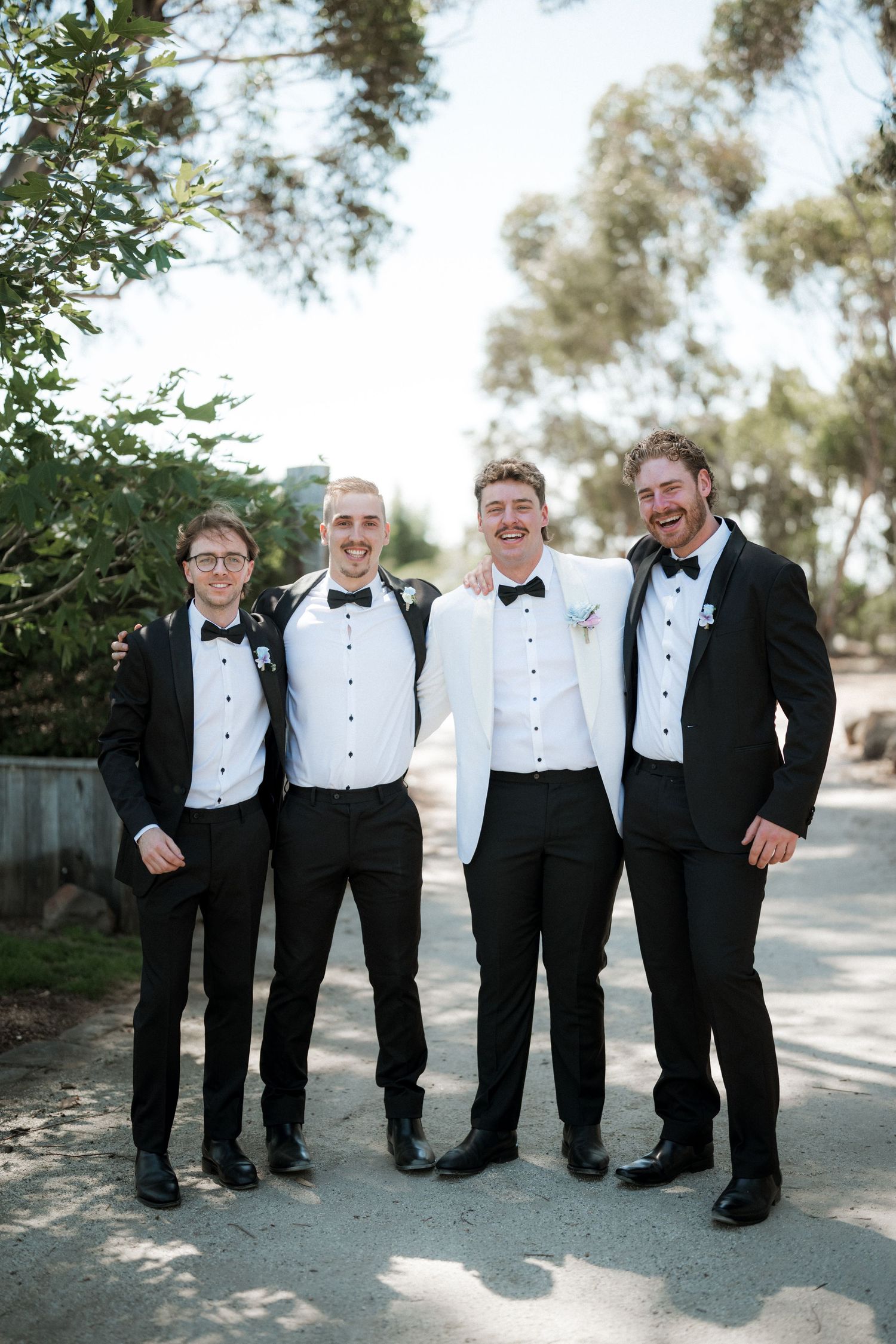 Groom in white jacket posing with three groomsmen in black tuxedos outdoors among trees.