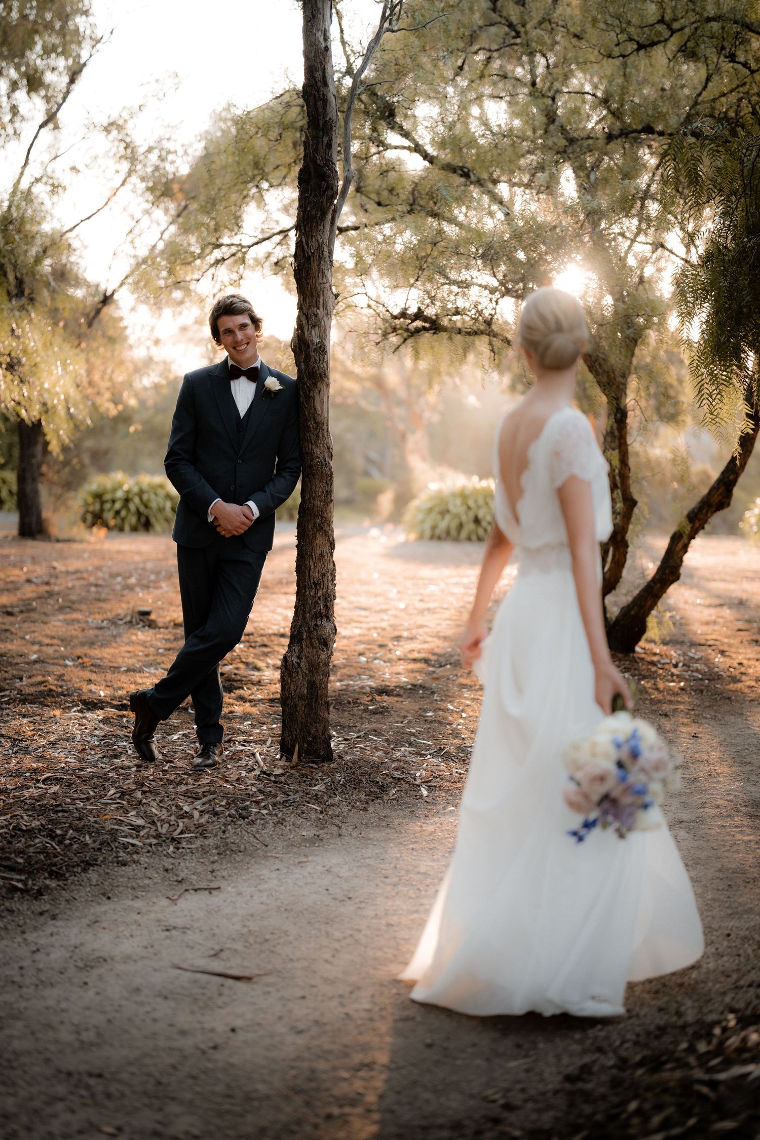 A romantic wedding photo in a wooded setting with sunlight filtering through trees during golden hour.
